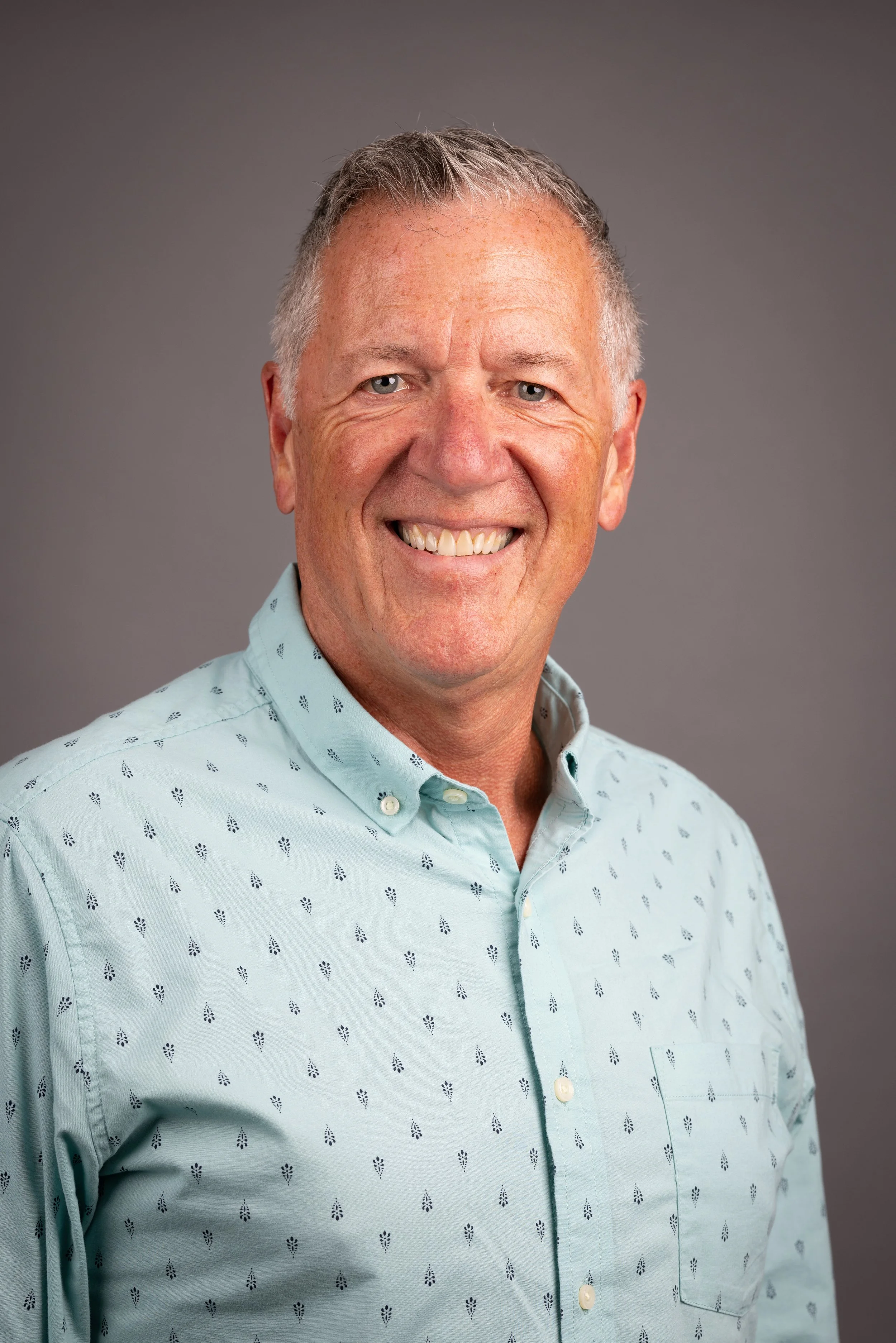 A smiling middle-aged man with short gray hair wearing a light blue button-up shirt against a gray background.