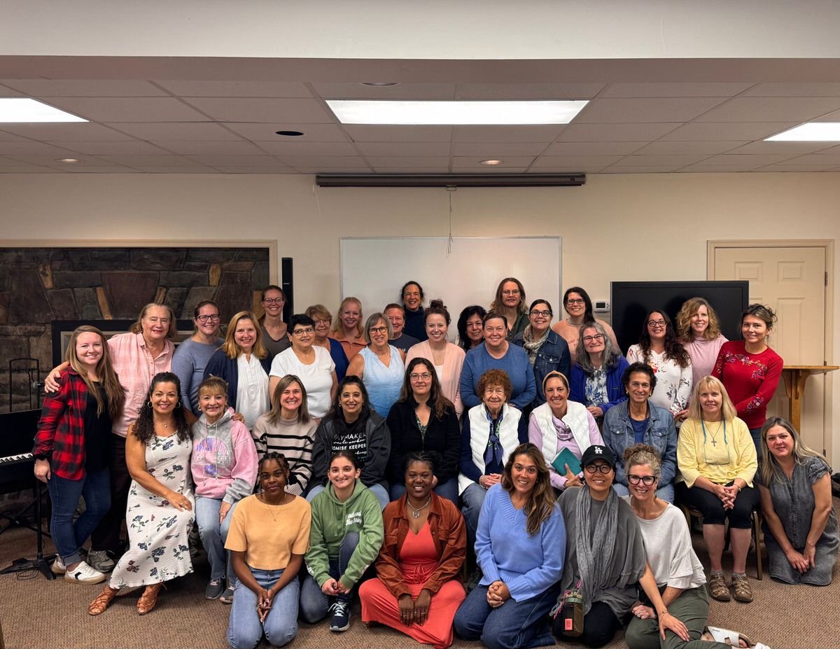 Group of diverse women gathered in a room for a photo, some seated and some standing, with a fireplace and a whiteboard behind them.
