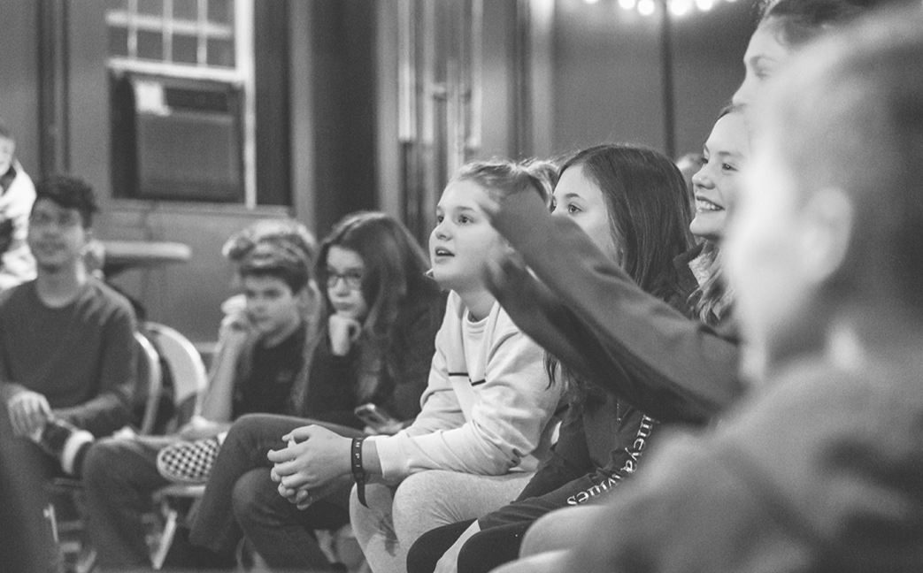 Group of children sitting in a room, attentively looking at something or someone outside the frame, some smiling and others with focused expressions.