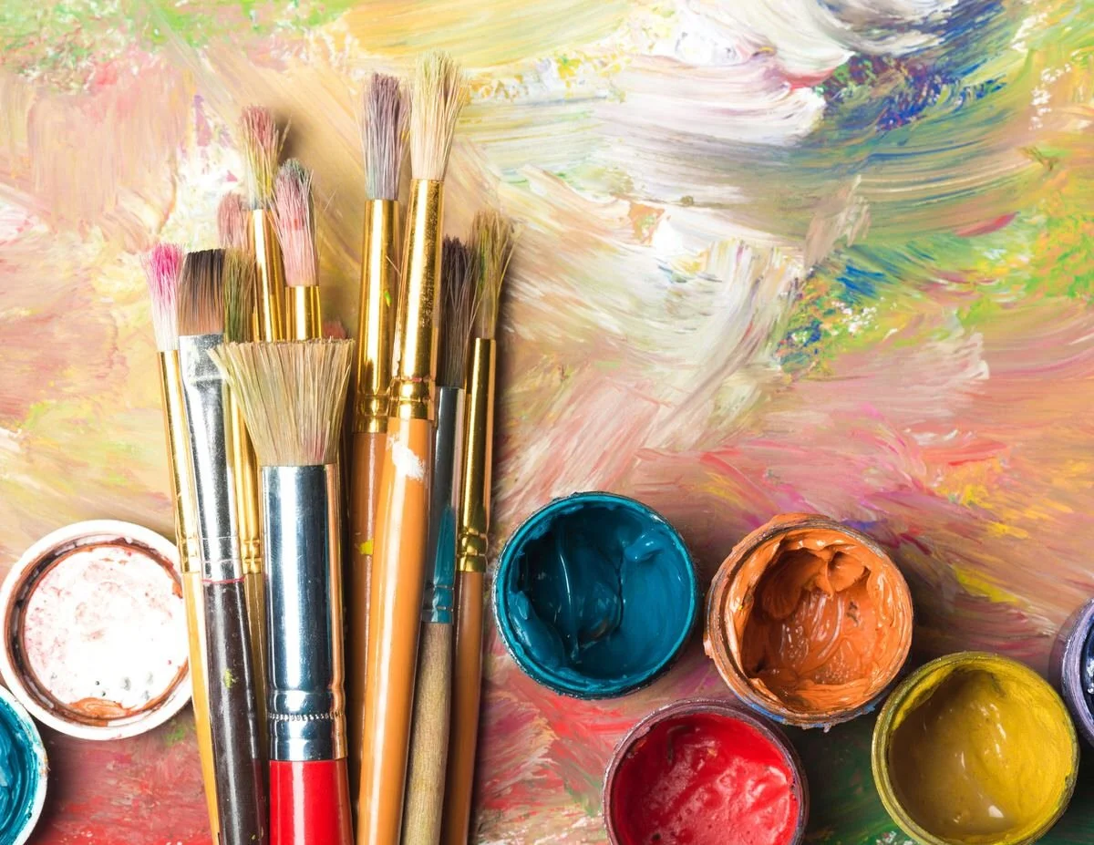 Paint brushes and open containers of blue, orange, red, and yellow paint on a multicolored, textured background.