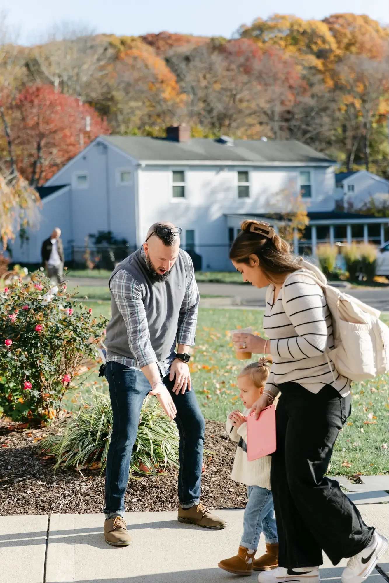A man appears to be showing or explaining something to a woman and a young girl on a sidewalk in a residential neighborhood, with a house and autumn trees in the background.