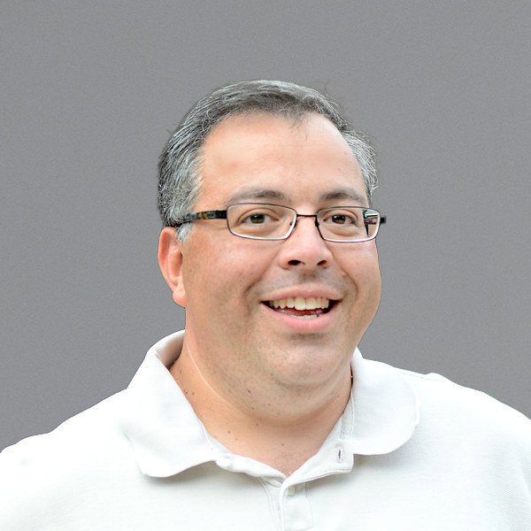 Headshot of a smiling man with glasses, beard, and short hair, wearing a black quilted jacket and a collared shirt, against a gray background.