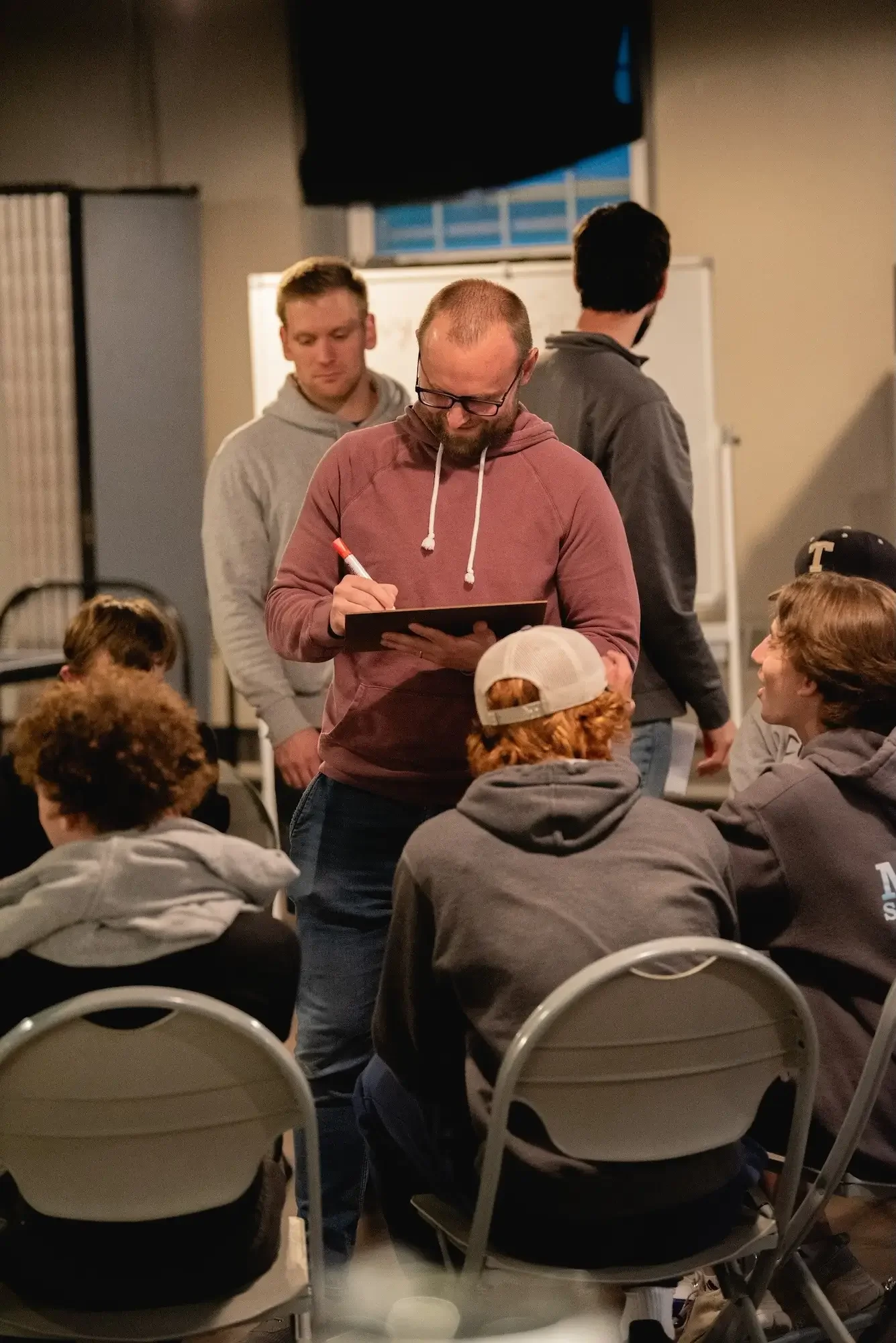 A group of young men in hoodies sitting in a room with a few standing men. One man in a maroon hoodie and glasses is writing on a clipboard. The room has chairs and a whiteboard.