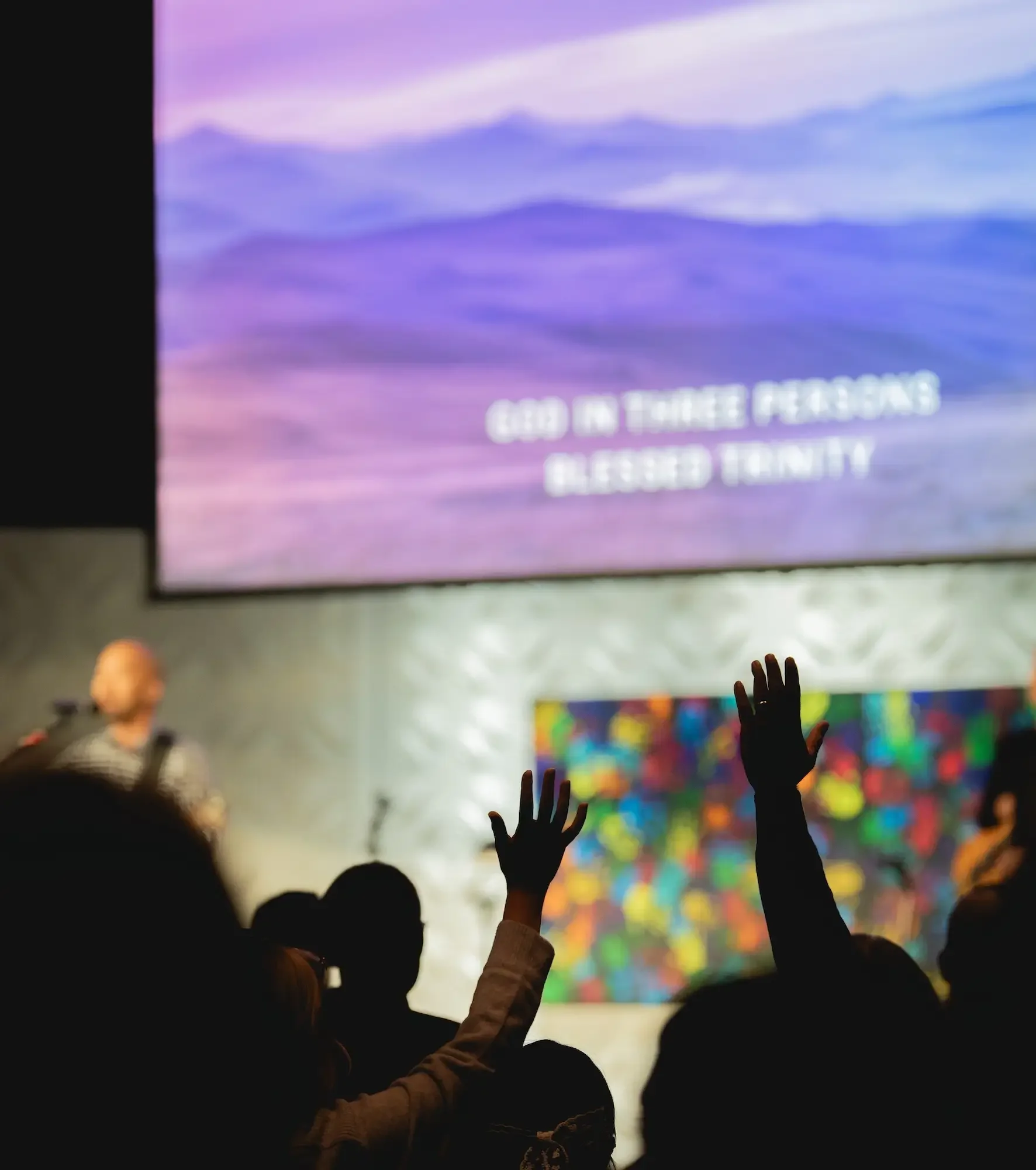 Crowd with raised hands attending a church service or worship event, with a large screen displaying the message "God in three persons blessed Trinity."