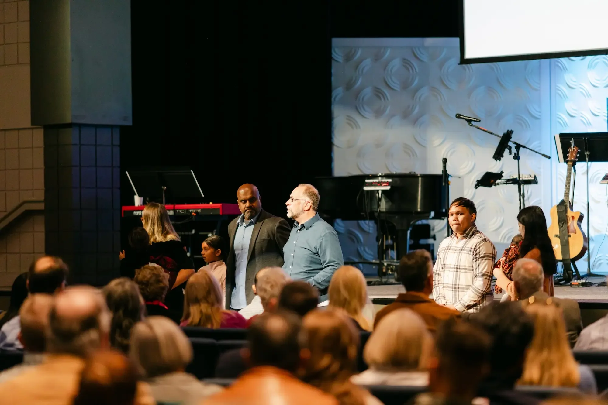 People standing on a stage in front of an audience, with musical instruments and microphones in the background.