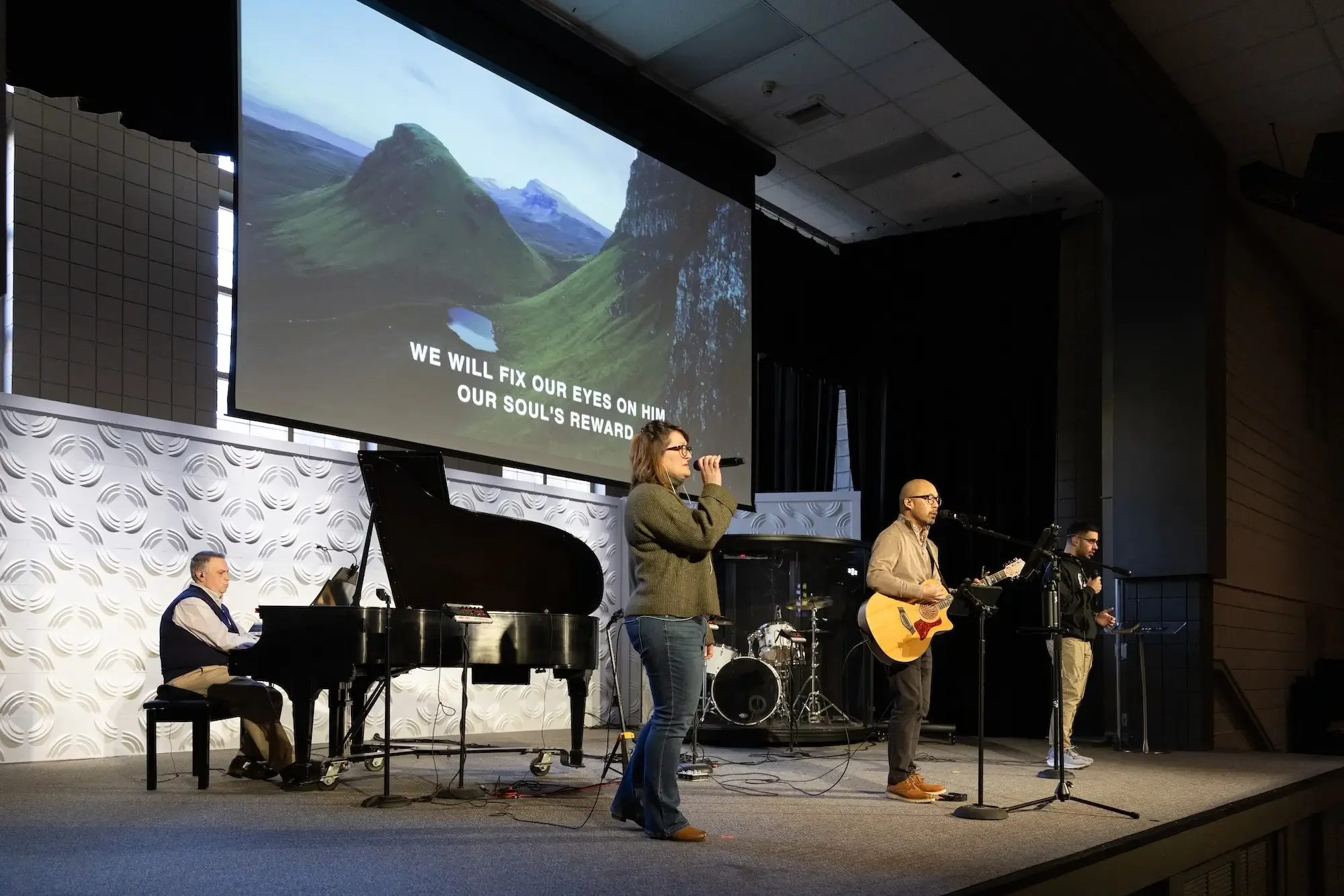 A band performing on stage with a pianist, a woman singing, and two men playing guitar and singing. A large screen in the background displays scenic mountains and a lake with the lyrics, "We will fix our eyes on Him, our soul's reward."