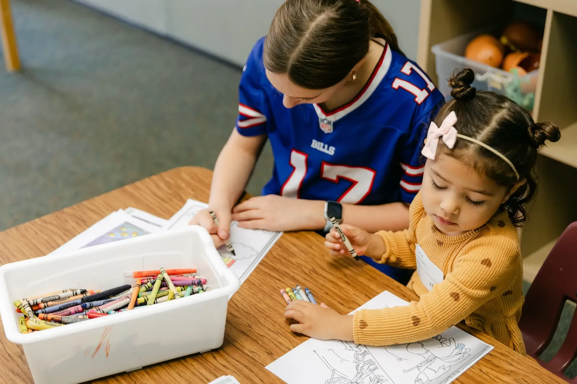 A woman and a young girl are sitting at a wooden table, coloring pictures with crayons. The woman is wearing a blue sports jersey, and the girl has curly hair with a pink bow, wearing a yellow sweater with heart patterns. drawing and coloring supplies are on the table and in a white container.