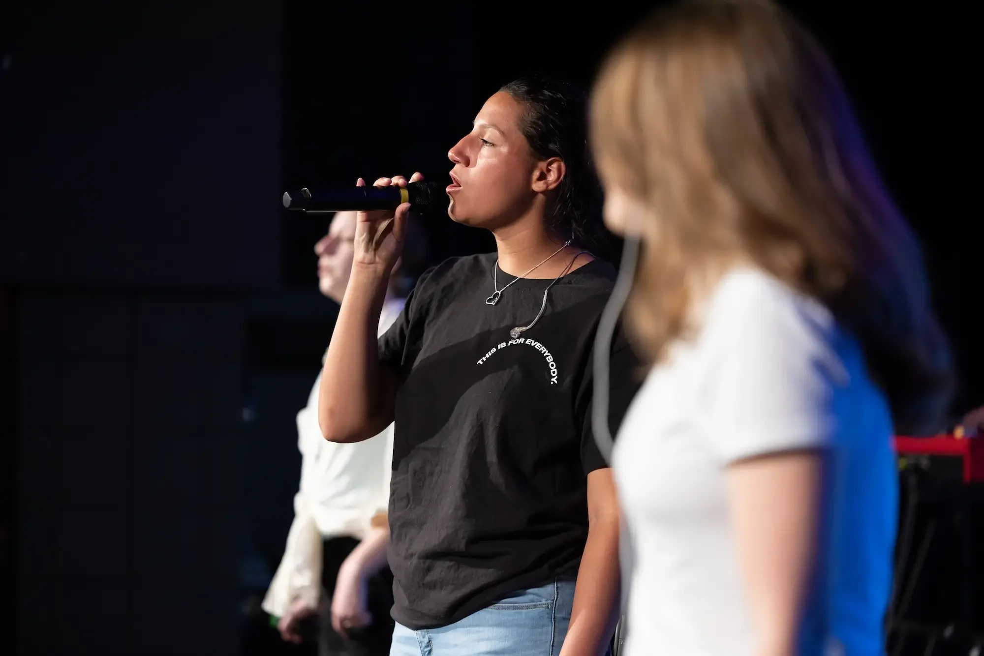 Young woman with dark hair holding a microphone during a stage performance, wearing a black t-shirt with white text, with other women blurred in the background.