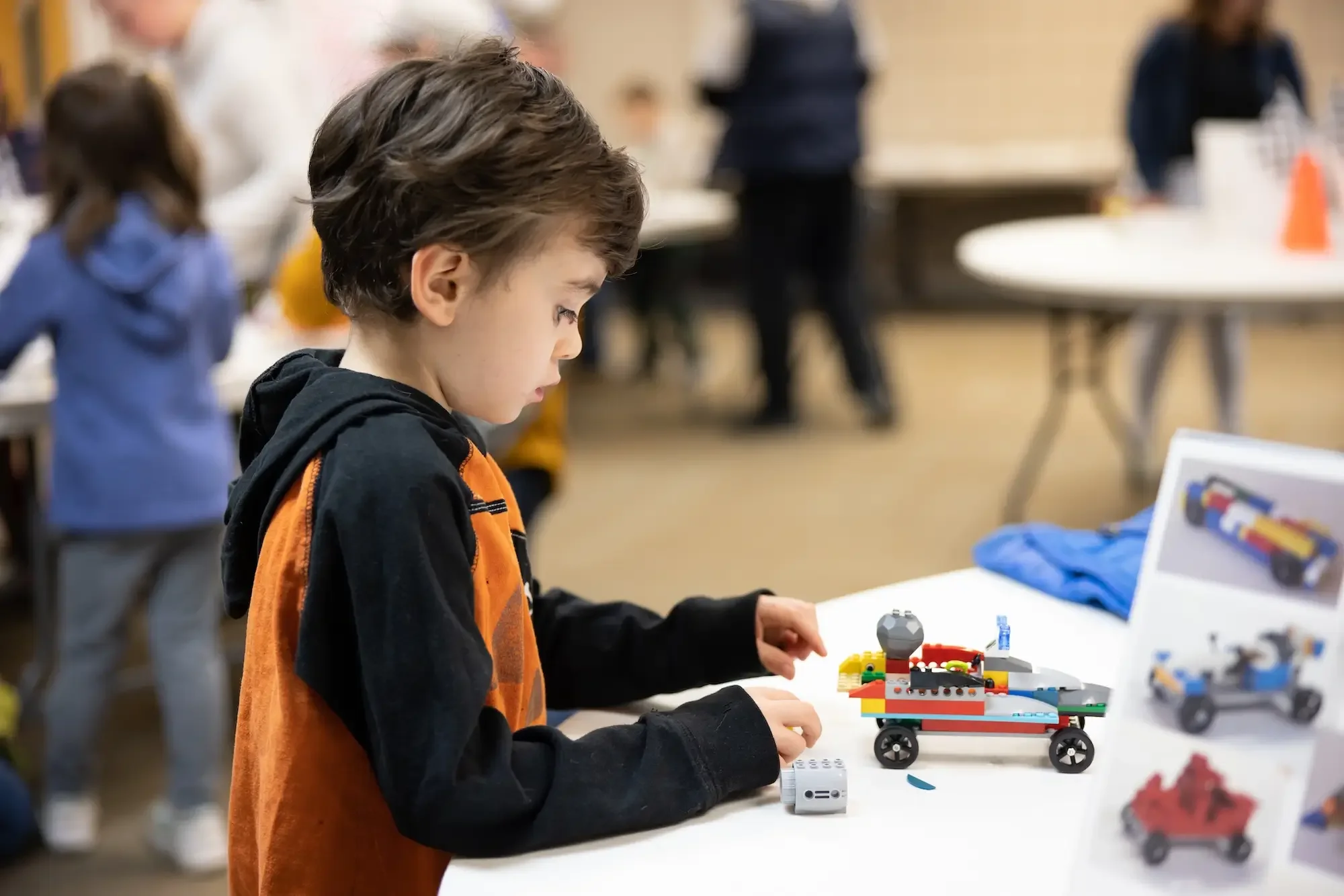 A young boy in a black and orange hoodie building a lego vehicle at a table during a robotics event.