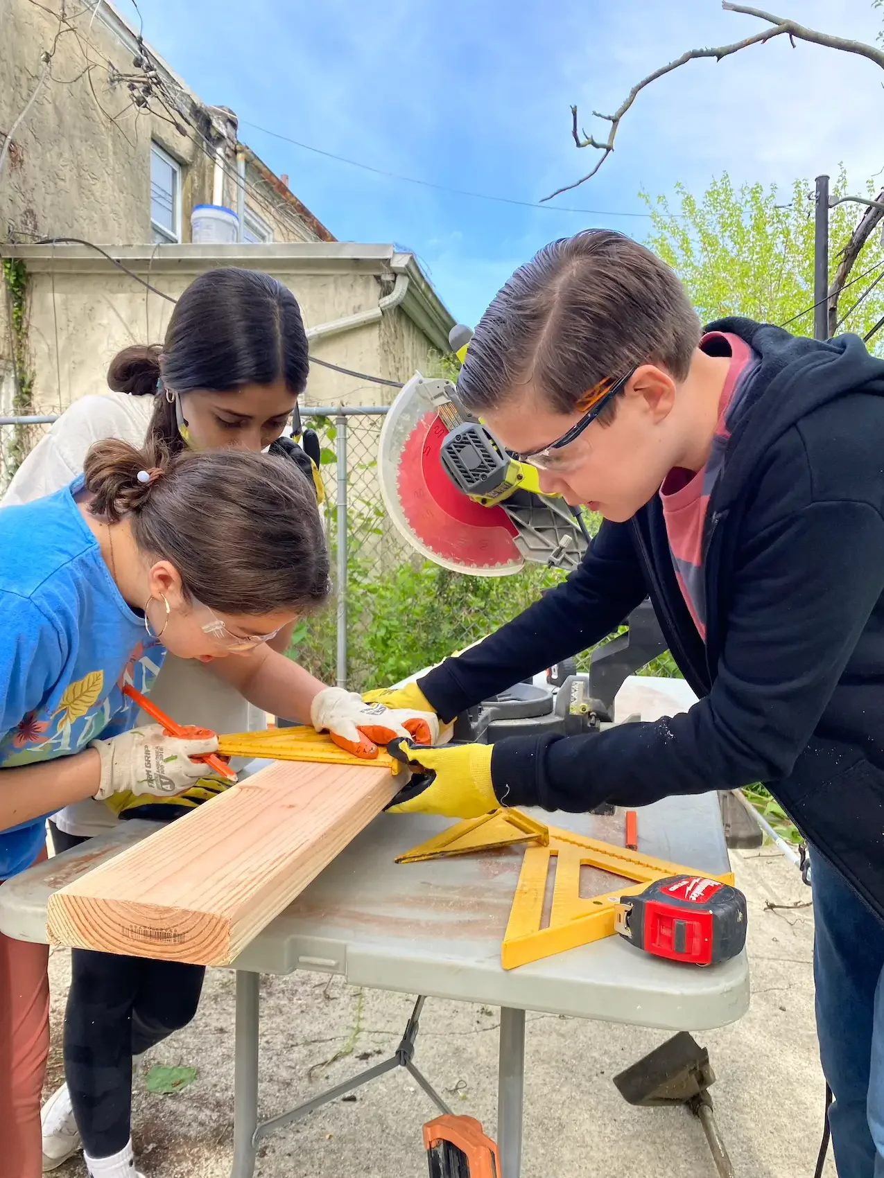 Group of four people working together on a woodworking project outdoors, using a measuring tape and saw on a wooden plank, during daylight.