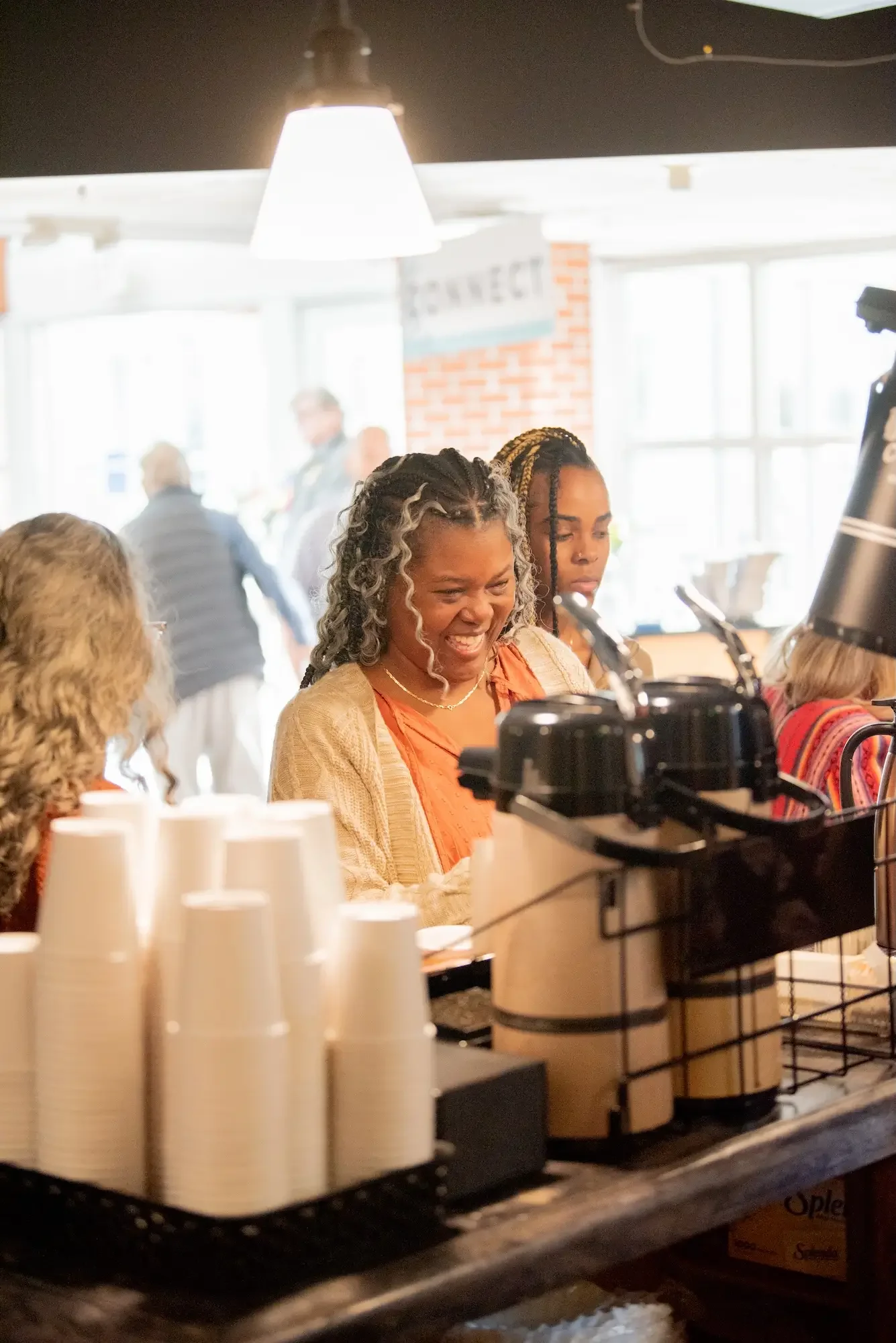Two women, one with curly gray hair and one with braided hair, smiling and enjoying their time at a coffee shop counter, with cups and coffee equipment in front of them.