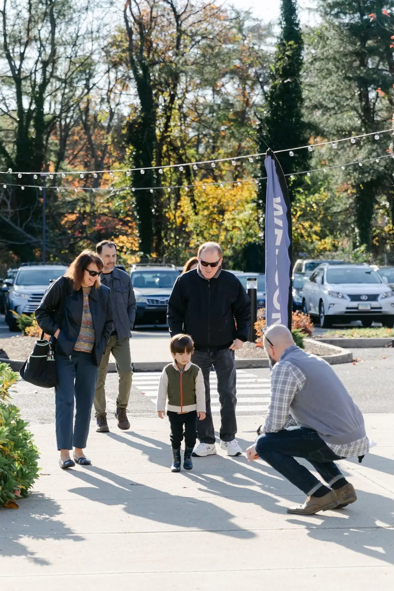A man crouching down and taking a photo of a young girl walking on a crosswalk, with four adults watching nearby on a sunny day in autumn, in a parking lot with trees in the background.