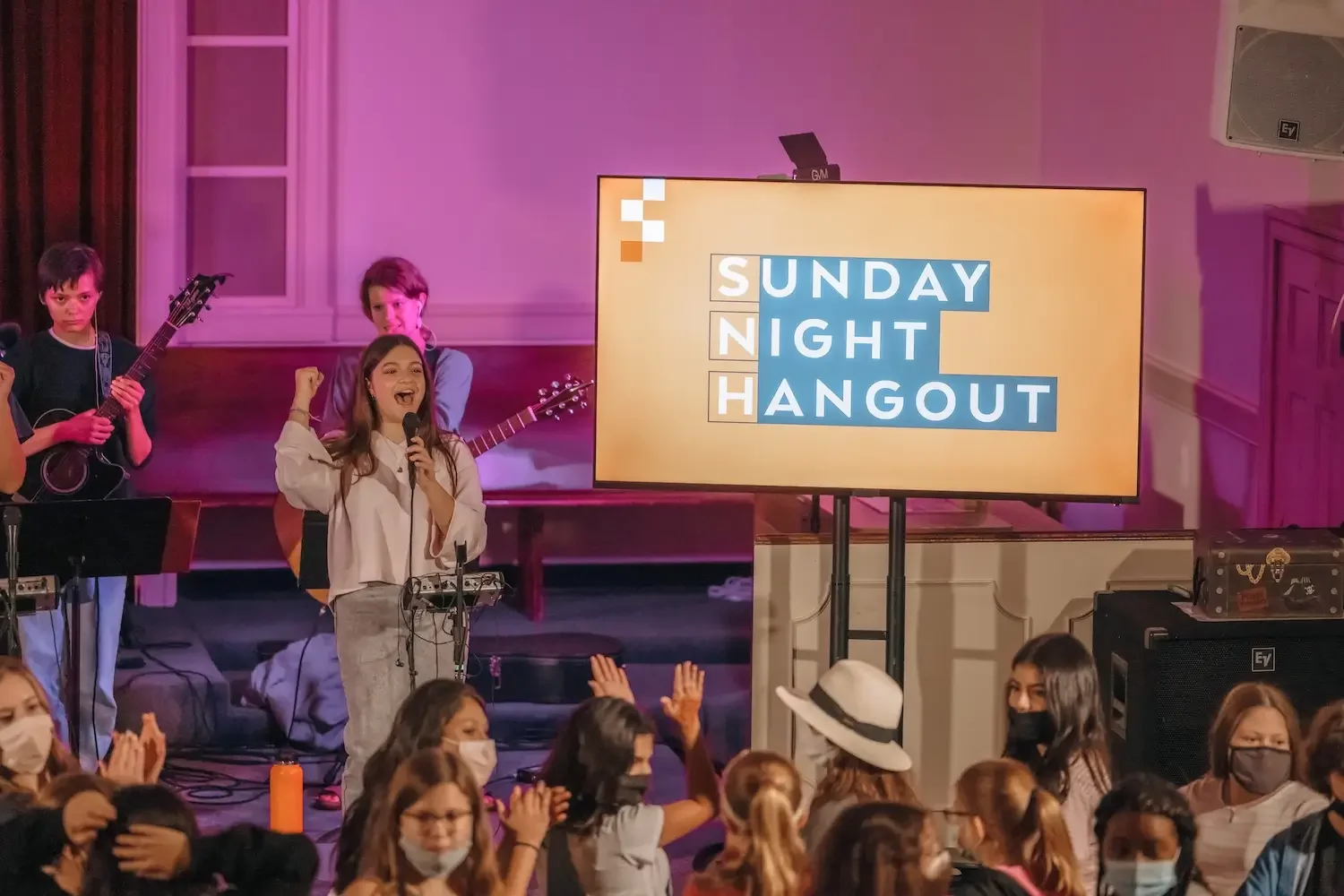 Young girl singing into a microphone with a band playing guitar behind her at a school event called Sunday Night Hangout; audience members wearing masks watching and clapping.