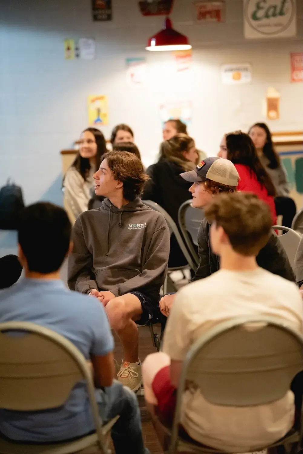 Group of teenagers and young adults gathering in a casual indoor setting, seated on chairs, engaging in conversation, with colorful posters and signs on the wall behind them.