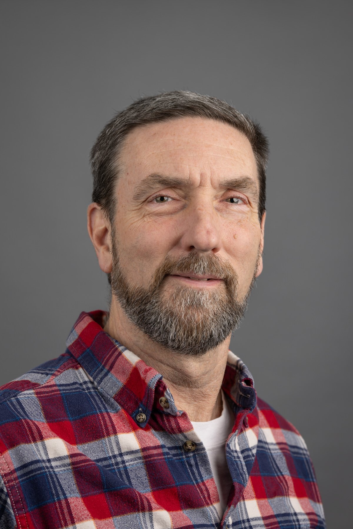 Headshot of a smiling middle-aged man with short light brown hair, wearing a pink button-up shirt against a plain gray background.