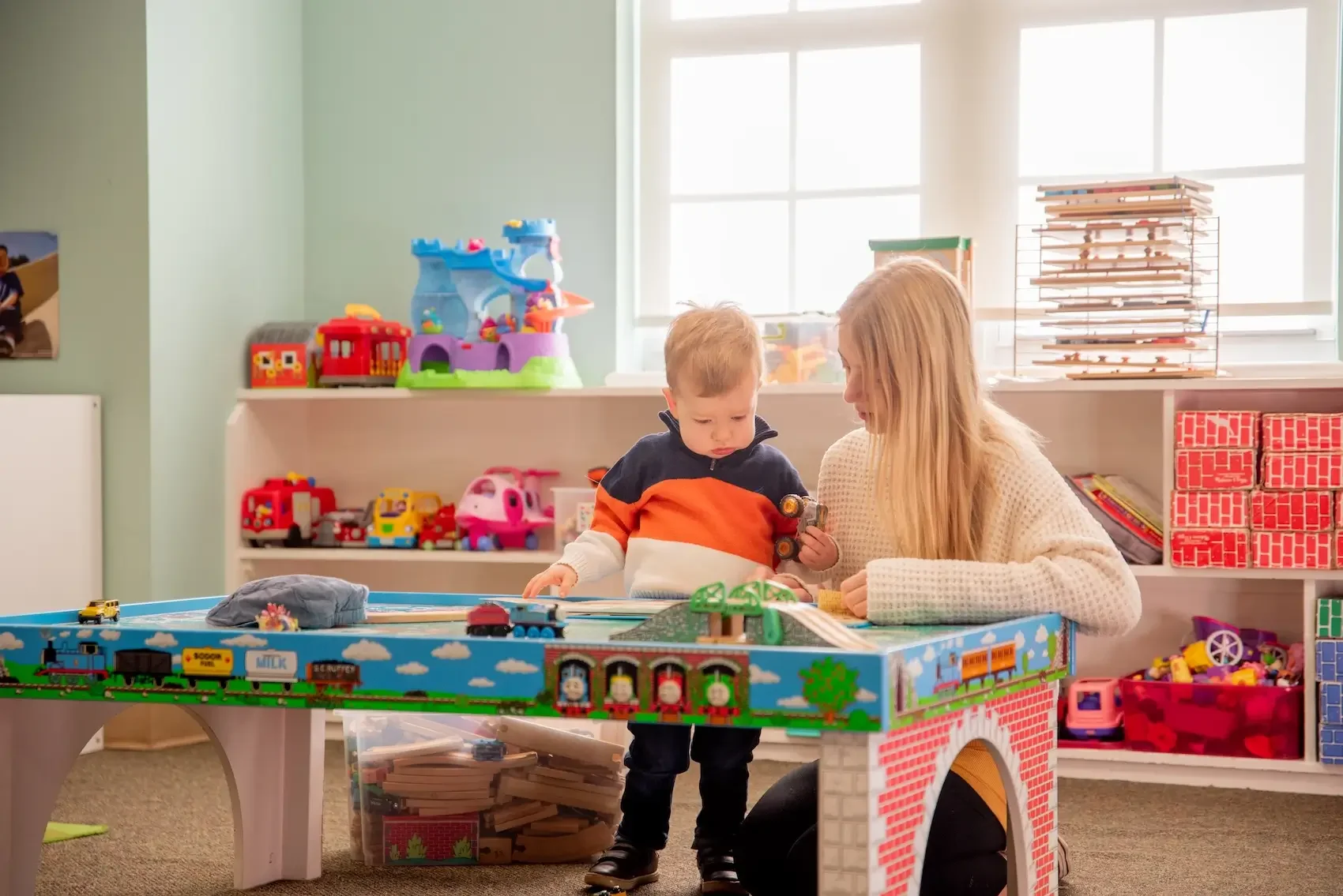 A woman and a young child playing with toy trains on a colorful train table in a playroom with shelves filled with toys and games.