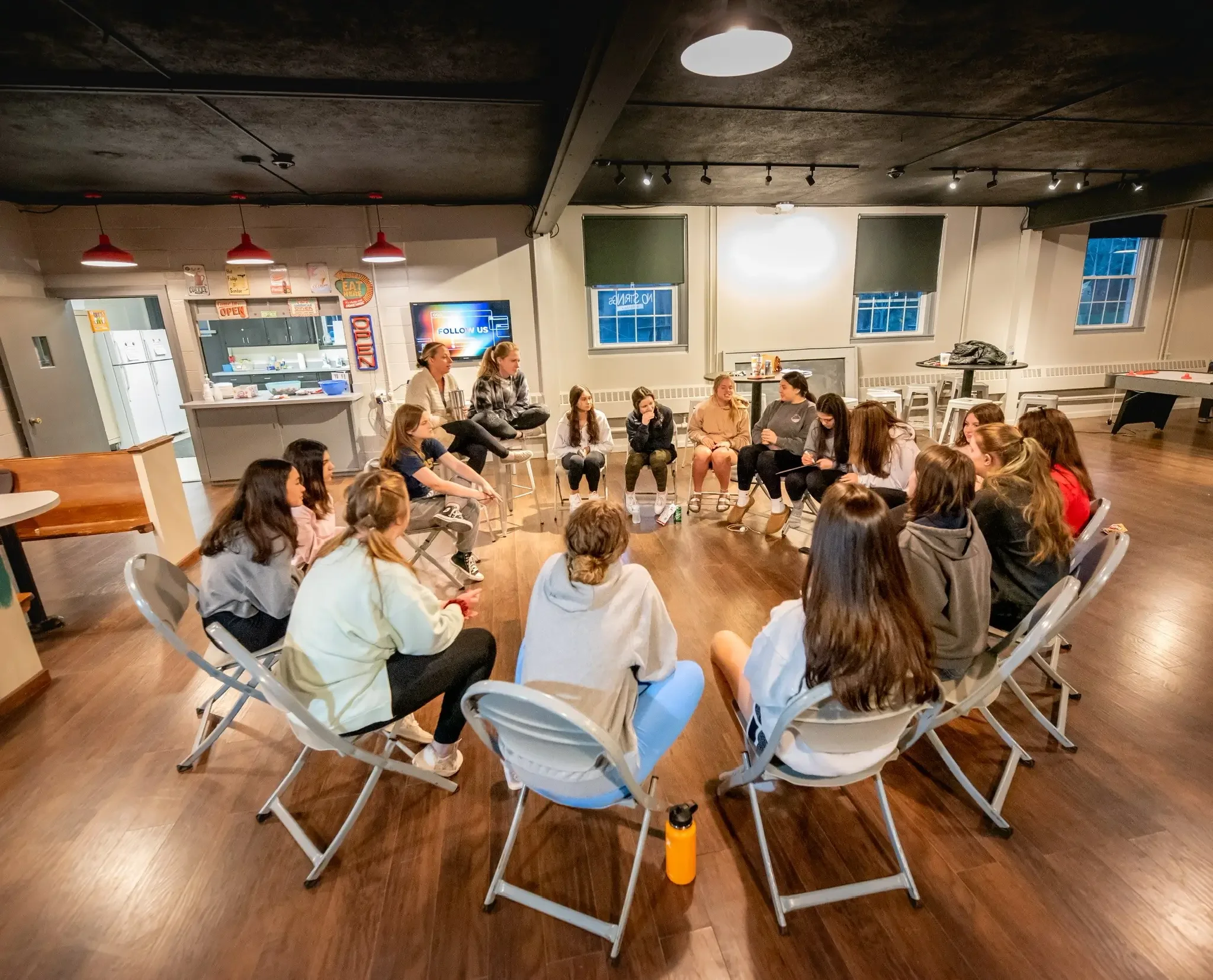 Group of young women and girls sitting in a circle in a room with wood floors, participating in a discussion or activity.