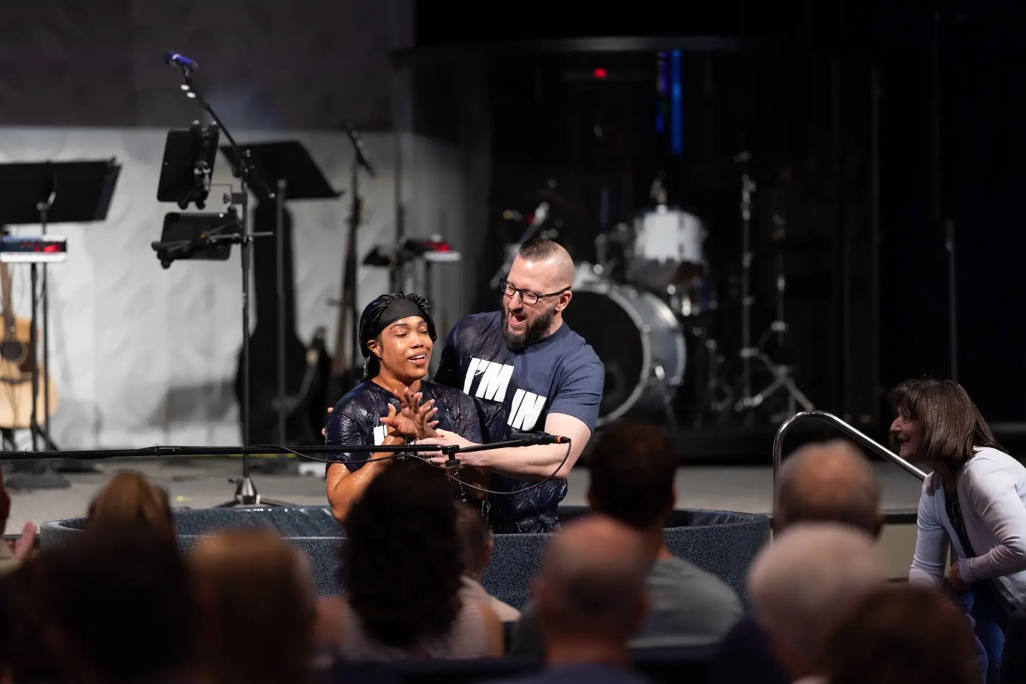 A man and woman on stage, with the man helping the woman out of a baptism pool during a church service, with audience members watching and smiling.
