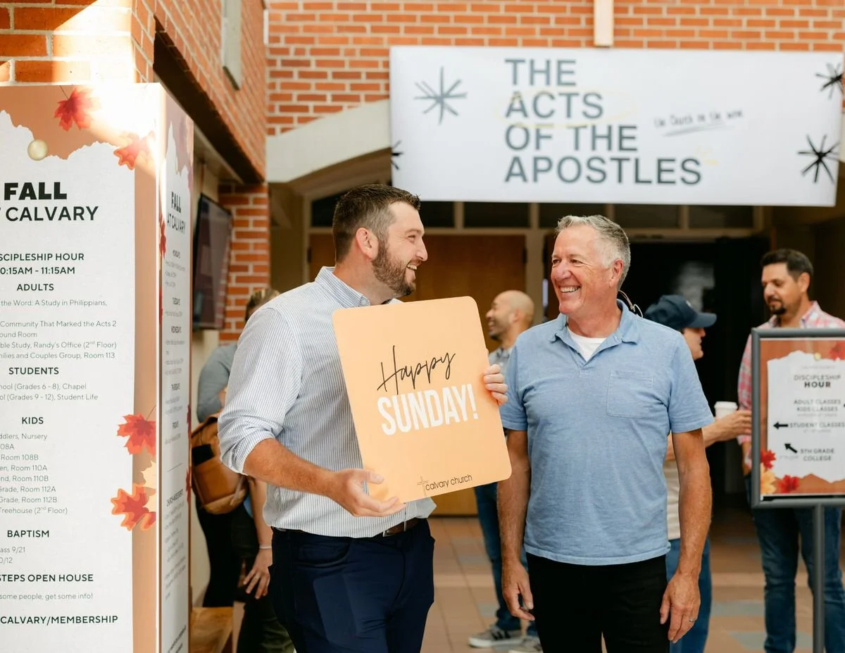 Two men smiling and talking at a church event, one holding a sign that says "Happy Sunday!" inside a church lobby with signs and people in the background.