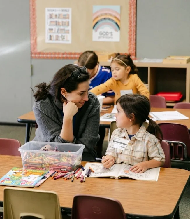 A classroom with a young girl and a woman sitting at a table, smiling and engaging with each other, surrounded by open books and colorful crayons. In the background, other children are working at their desks.
