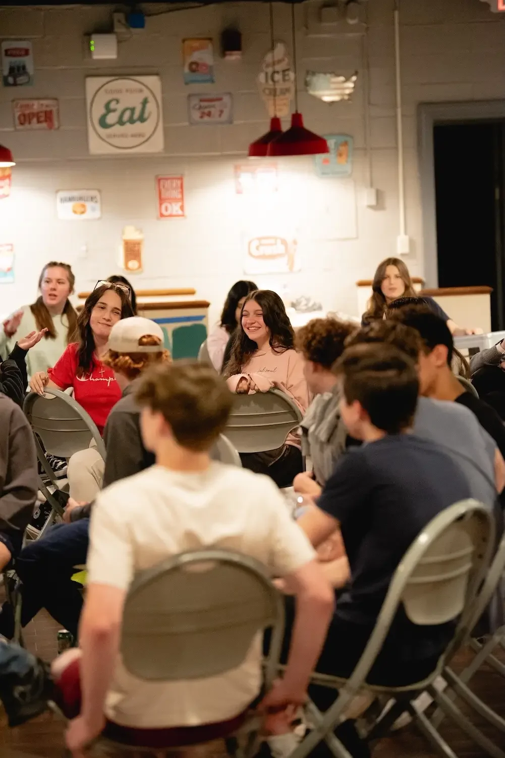 Group of teenagers sitting and talking in a casual setting, likely a cafe or restaurant, with posters on the wall and hanging red lamps. Some are smiling and engaging with each other.