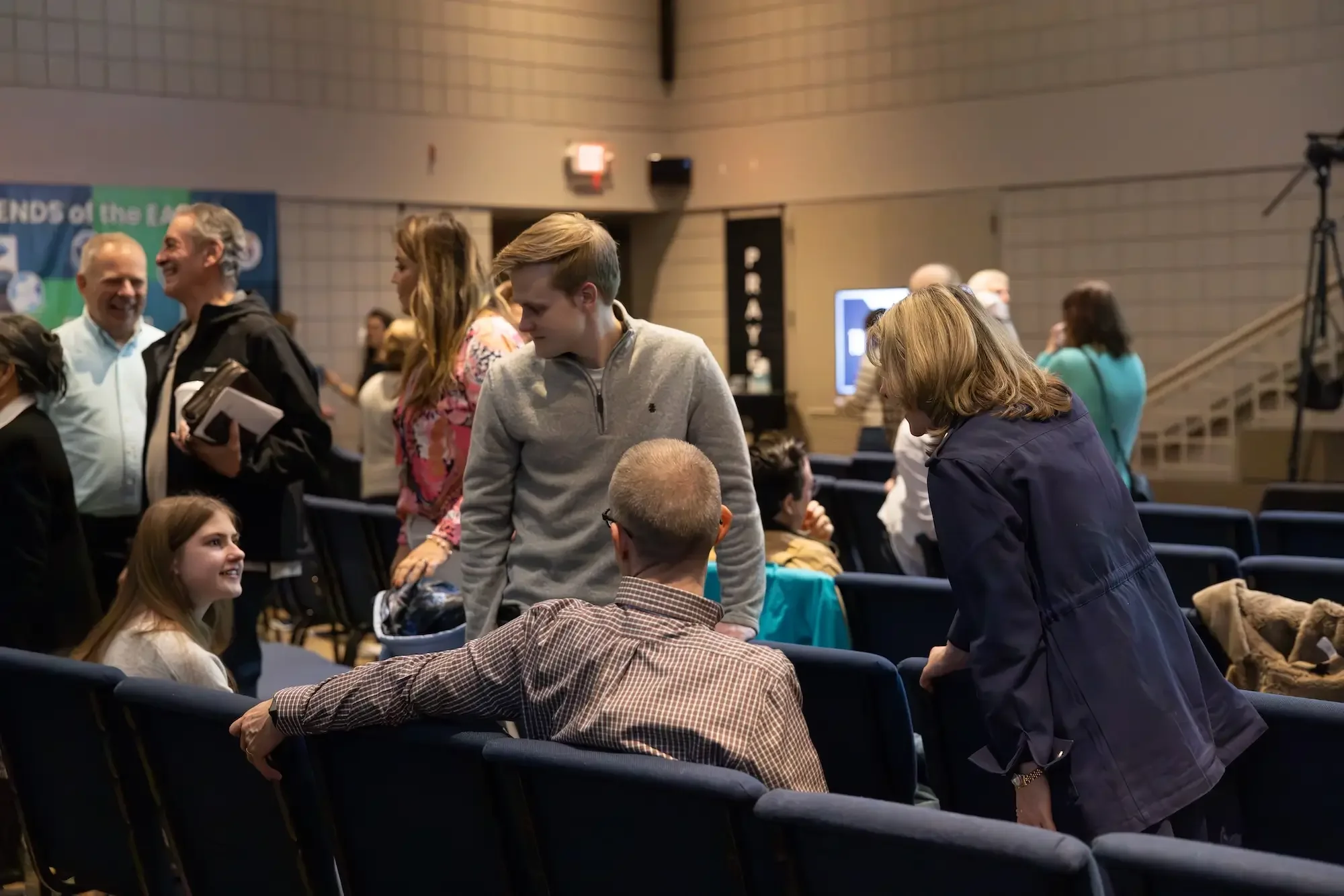 People gathered in an indoor auditorium, engaging in conversation and socializing, with rows of chairs, a digital screen, and a staircase visible in the background.