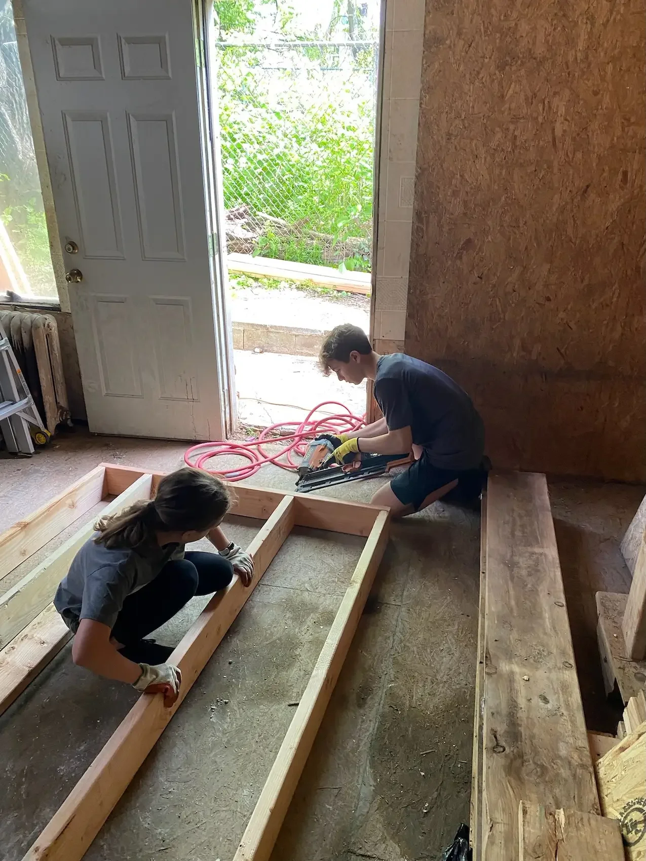 Two children building a wooden frame indoors, with a door open to a backyard. The children are using tools and wearing gloves, working on a construction project.