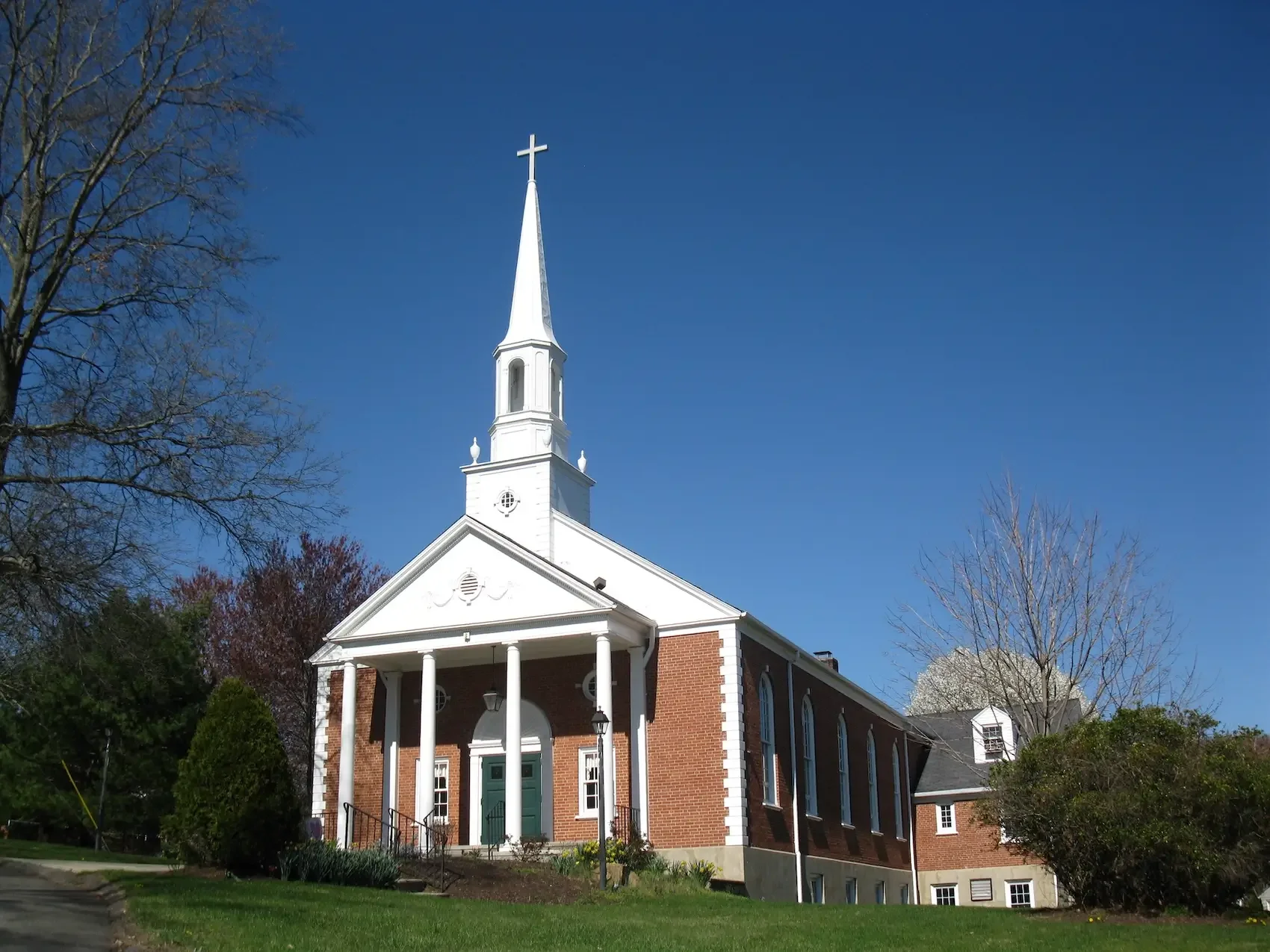 White church with steeple and cross, brick walls, green door, surrounded by trees and grass, clear blue sky.