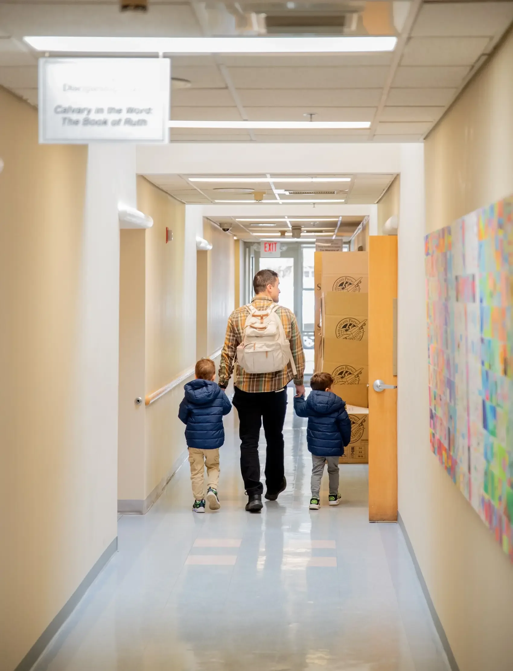 A man walking in a hallway with two young children, holding hands, heading toward the exit, with boxes stacked on one side and colorful artwork on the other.