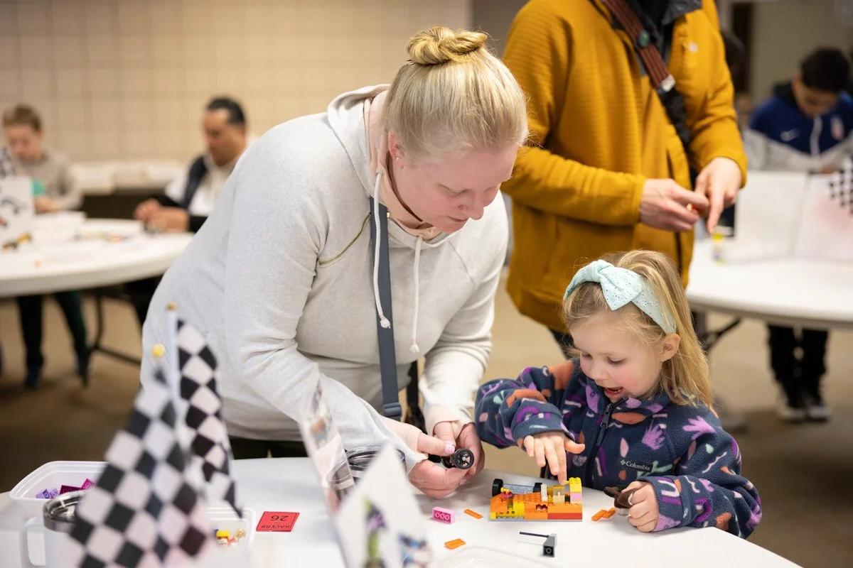 A group of young children and an adult woman playing with toys at a table in a classroom or playroom.