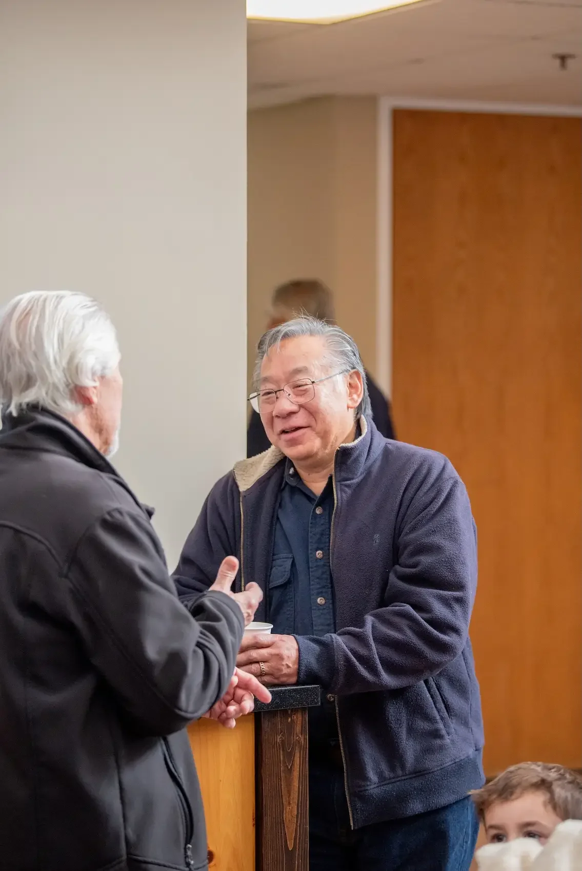 Two men engaged in conversation at an indoor gathering, one with white hair and a black jacket, the other with glasses and a navy jacket, standing near a corner with a child partially visible in the bottom right corner.