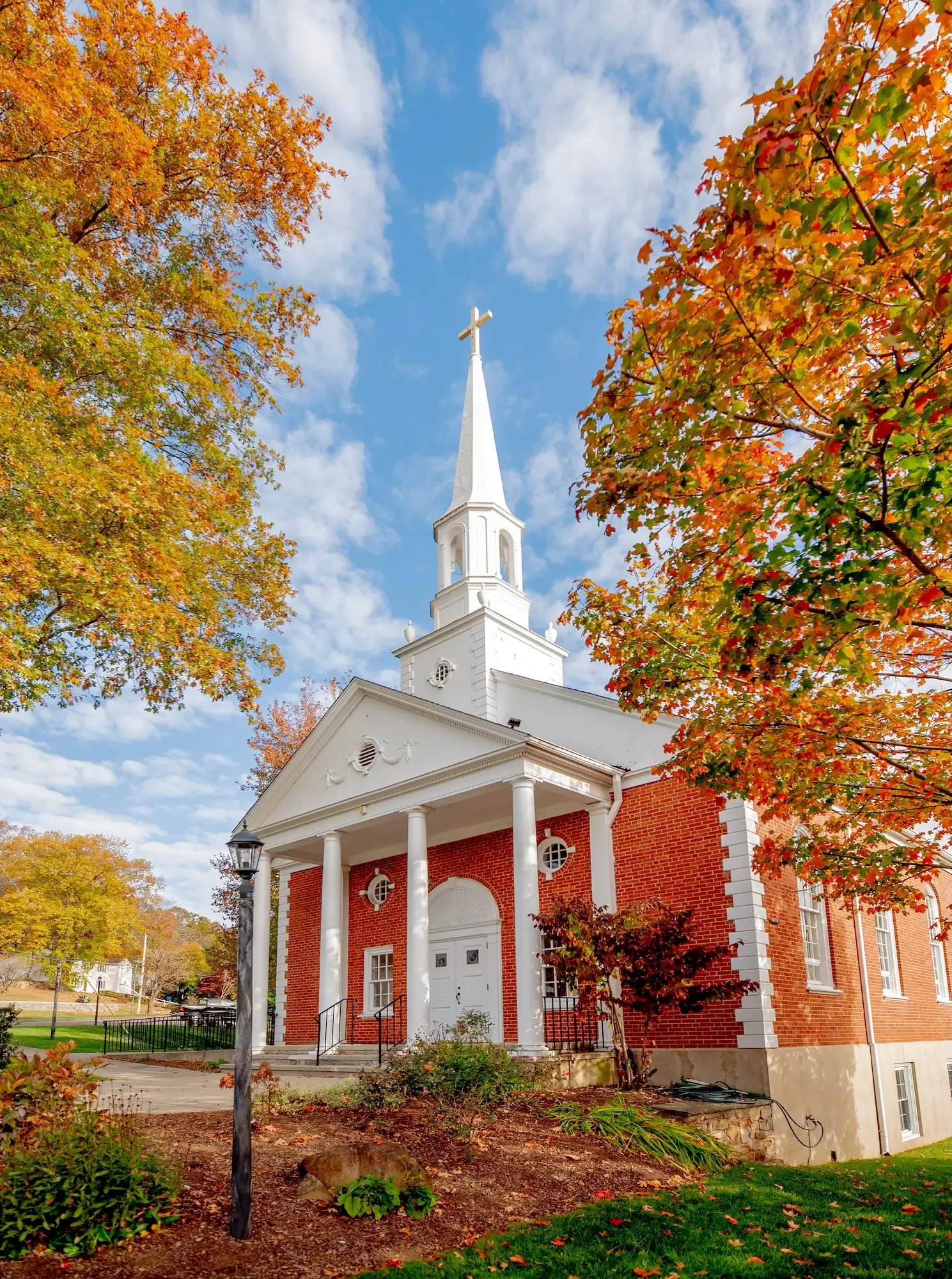 A white church with a tall steeple topped with a cross, surrounded by autumn trees with orange and green leaves, under a partly cloudy blue sky.