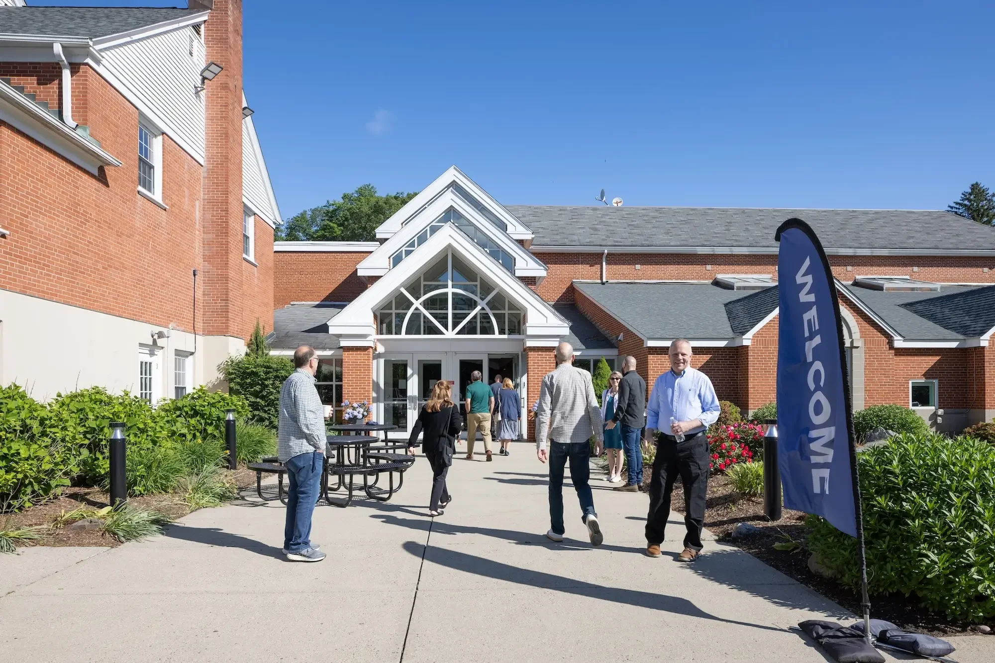 People gathering outside a brick building with large glass windows and a sign that says 'Welcome' on a flag. Clear sky and landscaped greenery visible.