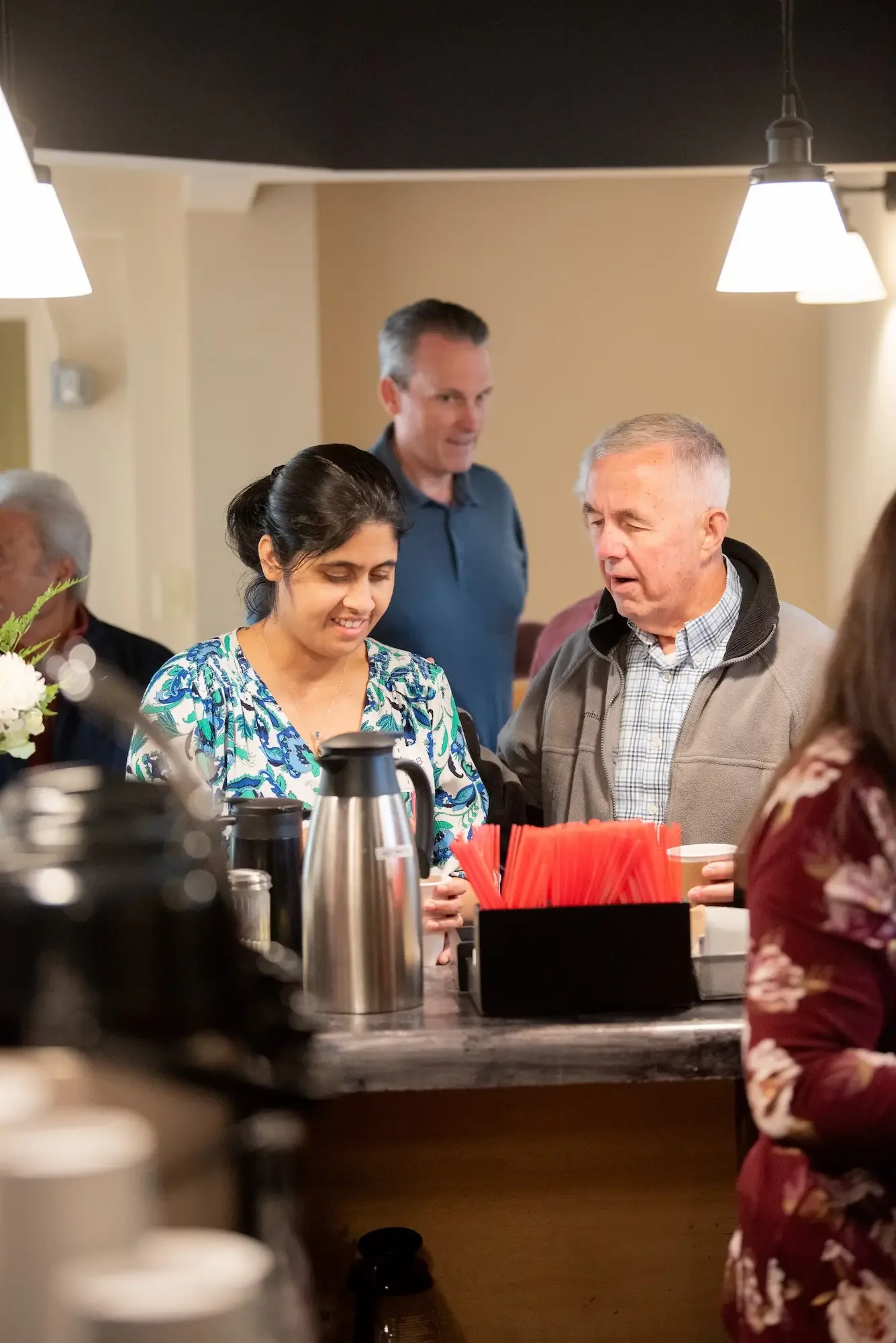People gathered at a coffee station, including a woman in a blue patterned shirt and an older man in a gray jacket, with another woman behind them and a man in a blue shirt in the background, in a cozy indoor setting.