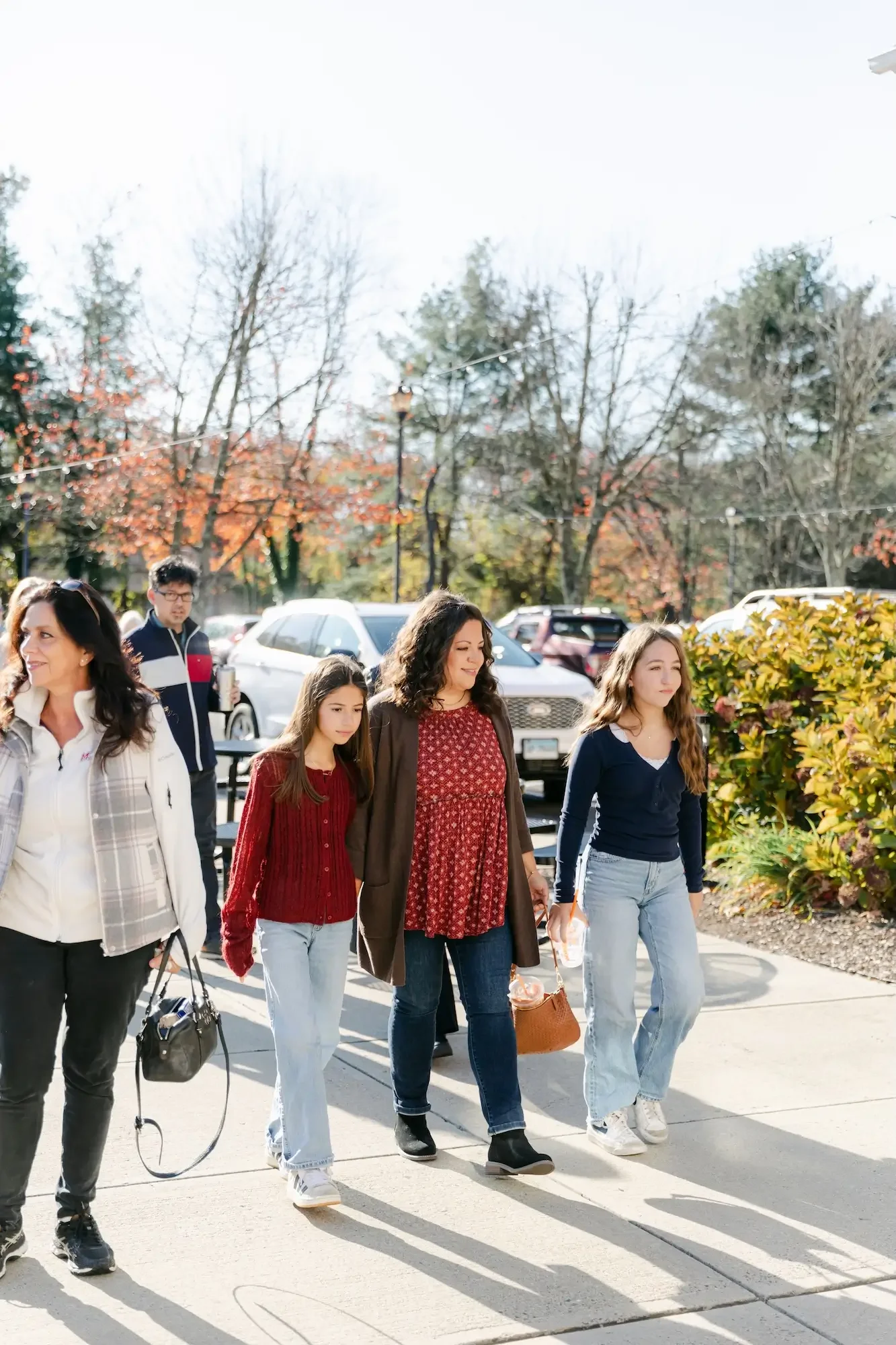Group of people walking outdoors in a parking lot area during daytime, with trees and parked cars in the background.