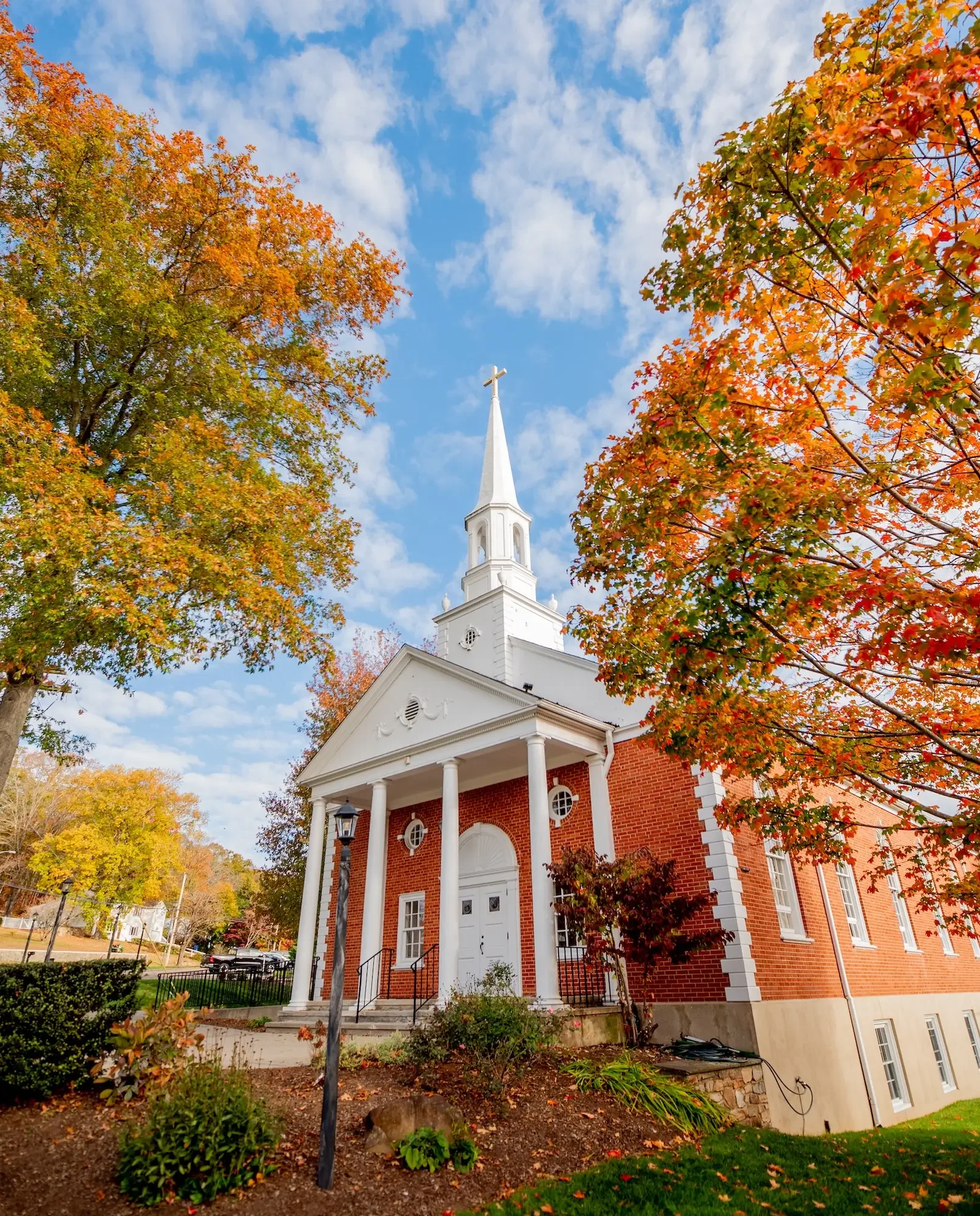A white church with a steeple and columns, surrounded by colorful autumn trees and a partly cloudy sky.