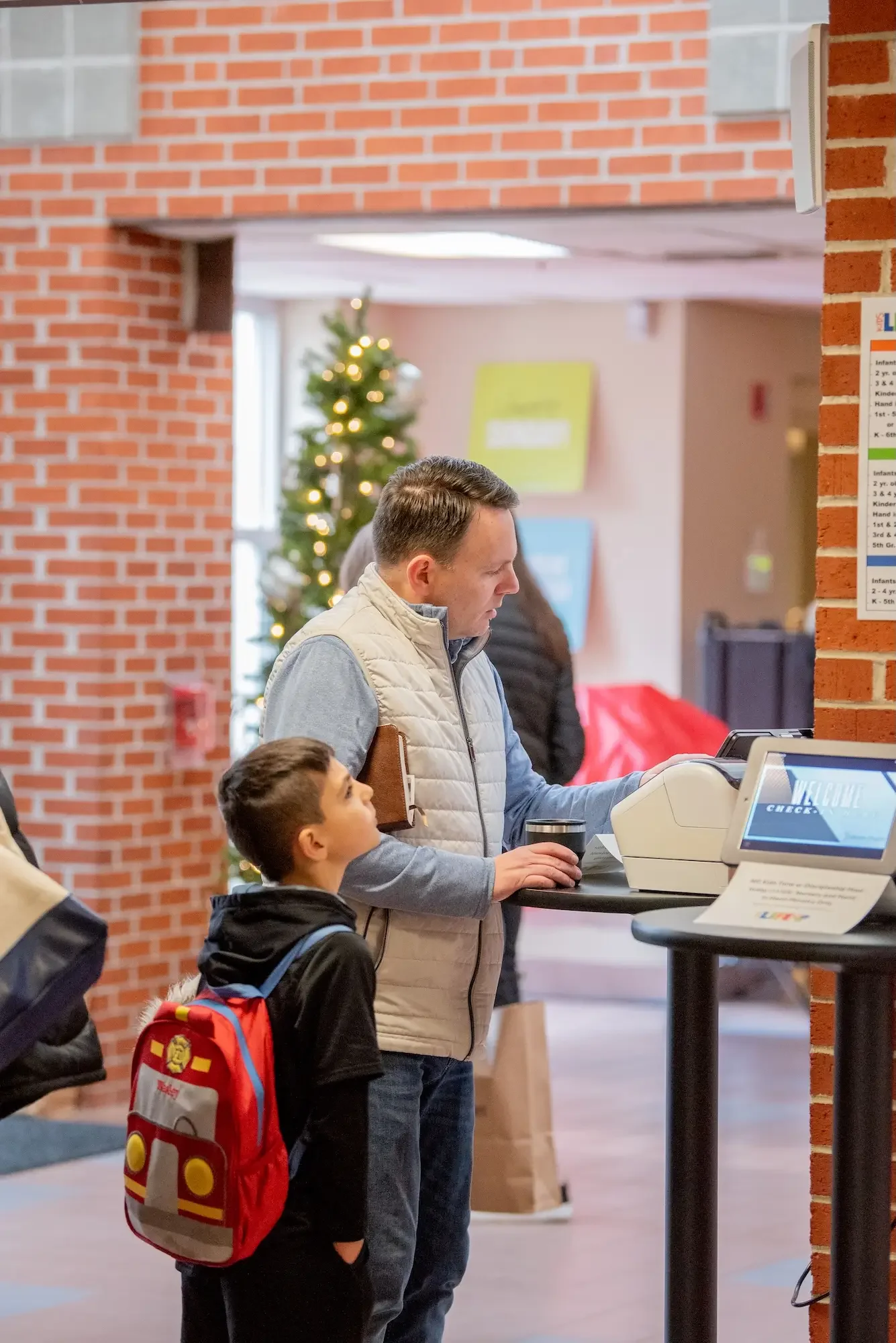 A man and a boy stand in line at a lobby counter, with a decorated Christmas tree with lights in the background.