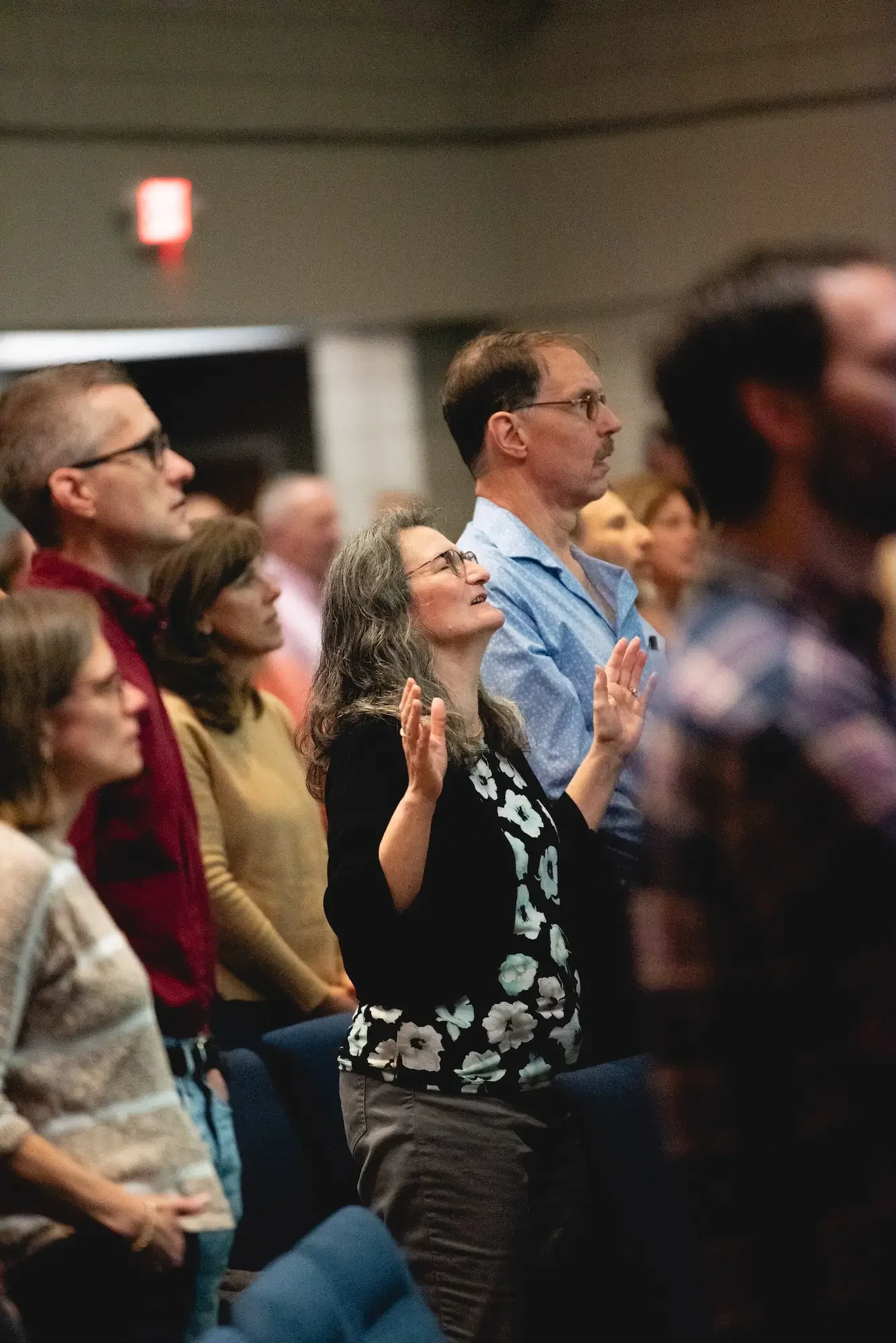 A group of diverse people standing in a congregation, some with their eyes closed and hands raised, participating in a religious service or prayer.