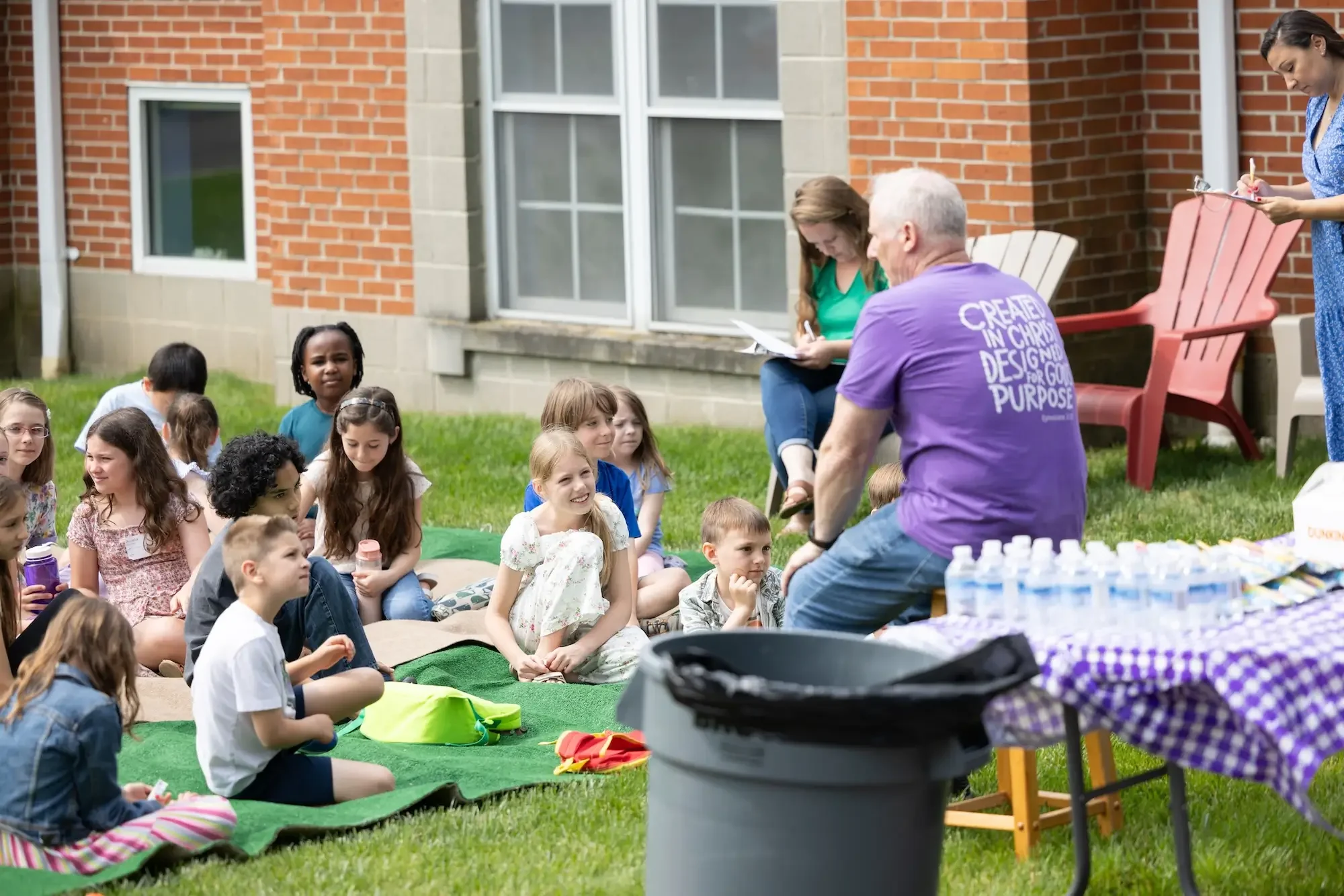 Group of children sitting on the grass listening to a man speaking outdoors during a gathering or event, with adults sitting nearby and bottles of water on a table.