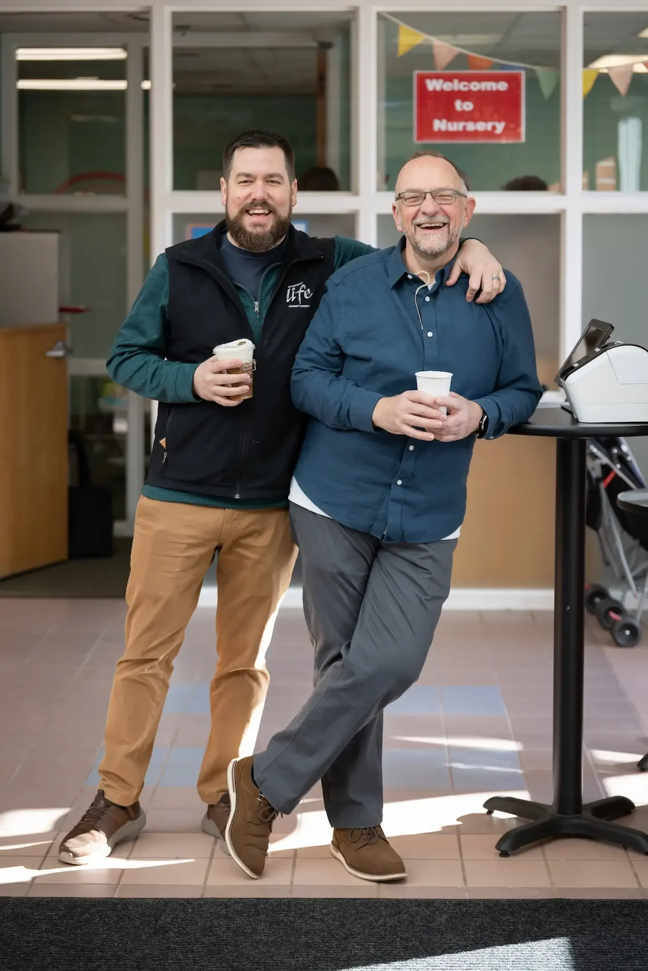 Two men smiling and holding cups of beverage inside a daycare or nursery, with a 'Welcome to Nursery' sign in the background.