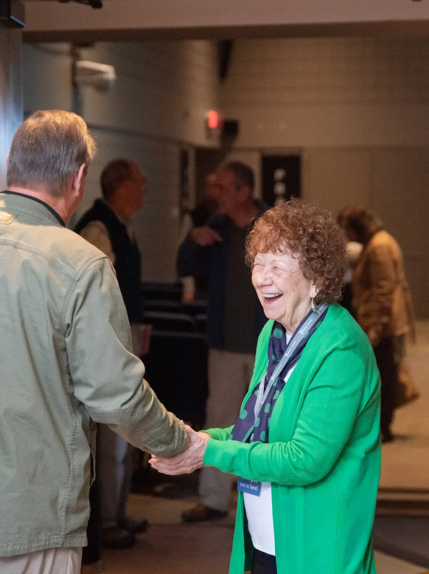 An elderly woman with curly hair, wearing a green cardigan and a polka-dotted scarf, is smiling and shaking hands with a man in a beige jacket in a social gathering or event.