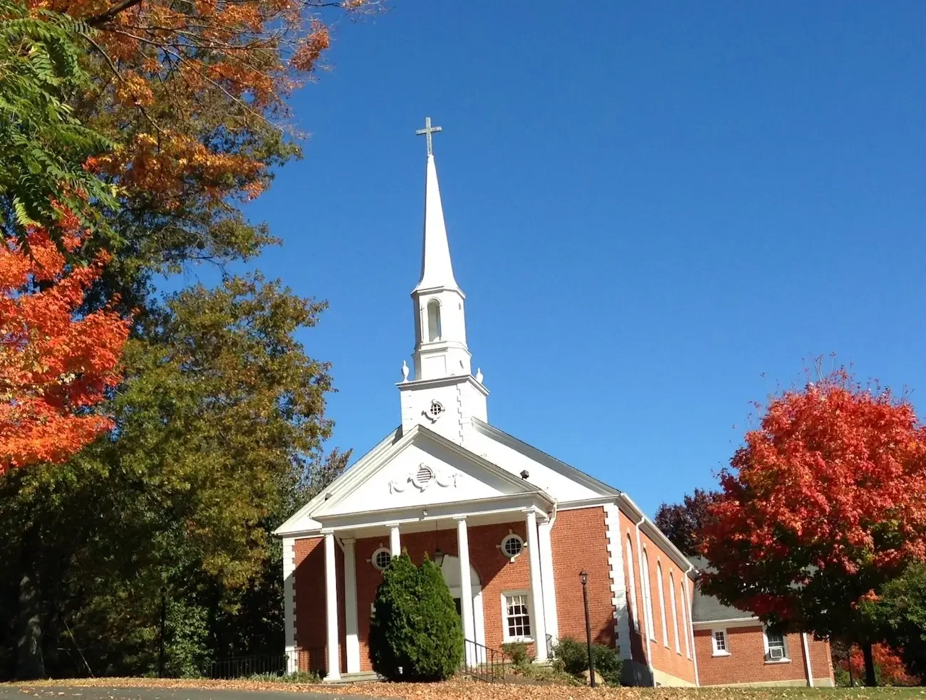 A white church with a tall steeple topped by a cross, surrounded by autumn trees with red and orange leaves, under a clear blue sky.
