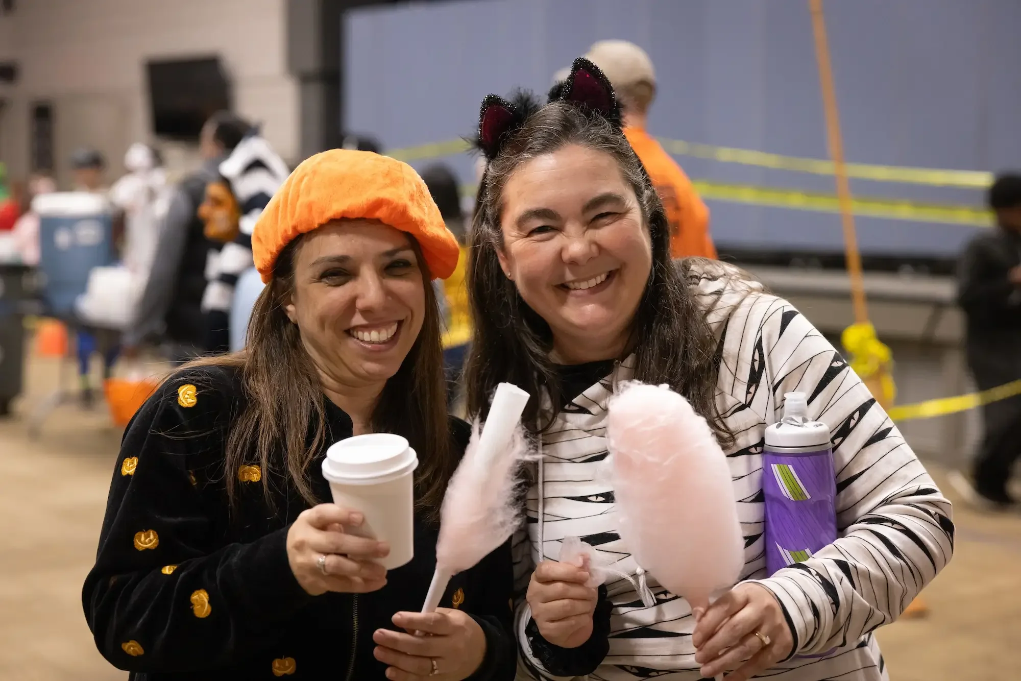 Two women smiling indoors, dressed in costumes, holding cotton candy, wearing Halloween-themed accessories, with a background of other people and event setup.