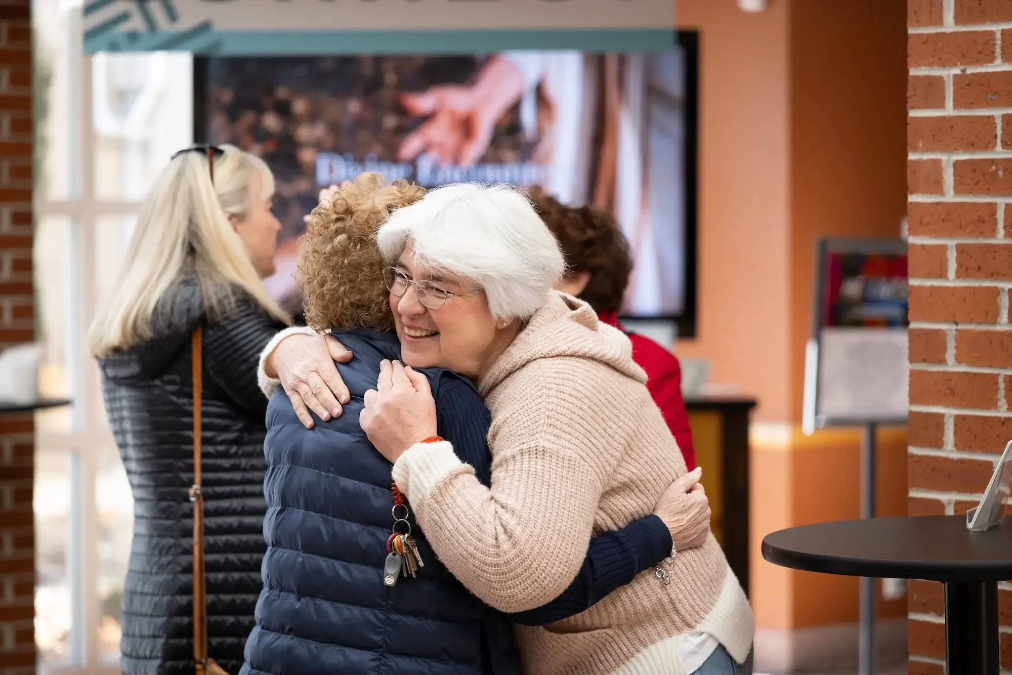 Two elderly women hugging and smiling at each other in a warmly lit indoor space, with other people in the background.
