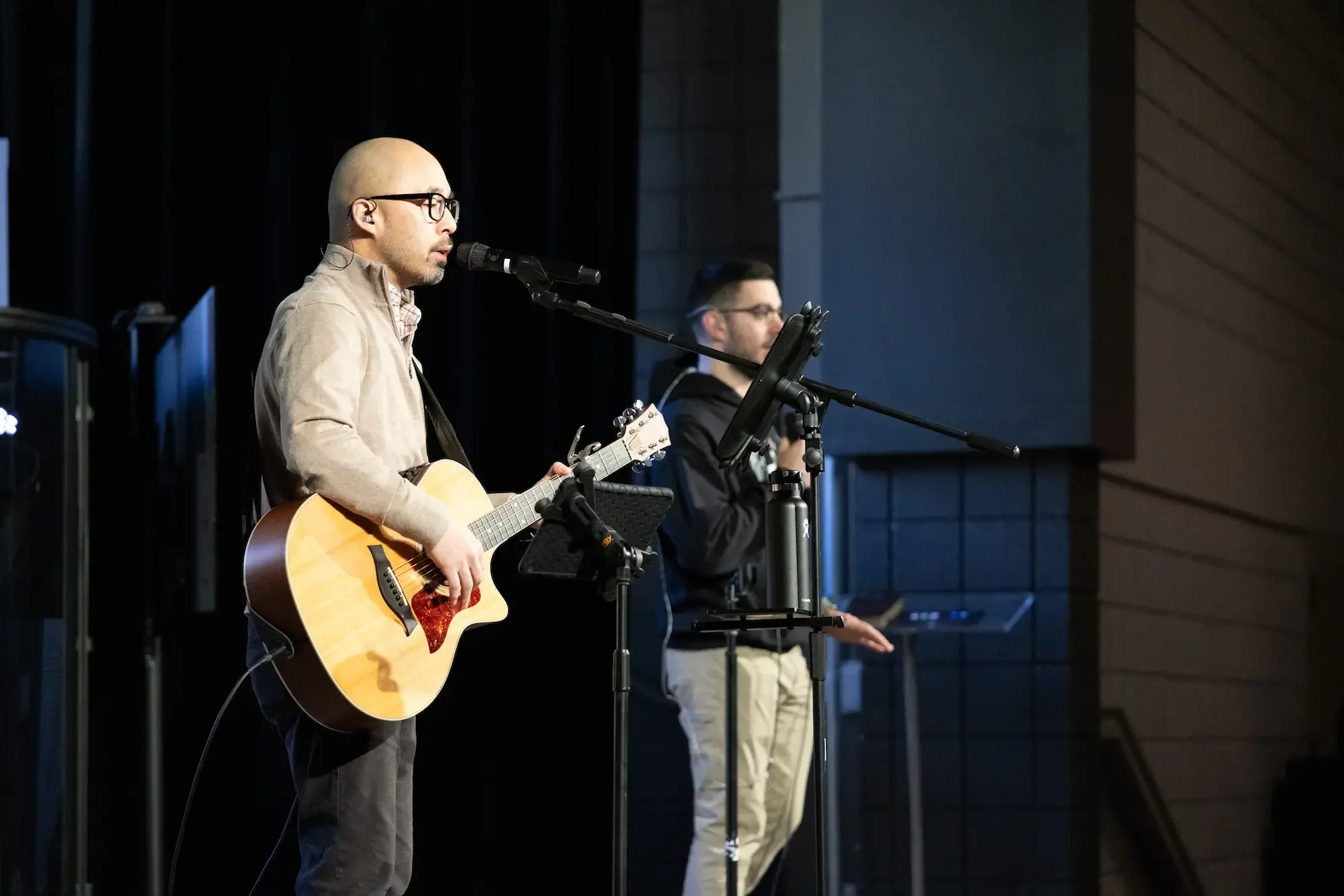 Two men on a stage, one playing an acoustic guitar and singing into a microphone, the other standing with a device in hand, in front of music stands and equipment, against a background of black curtains and a wall.