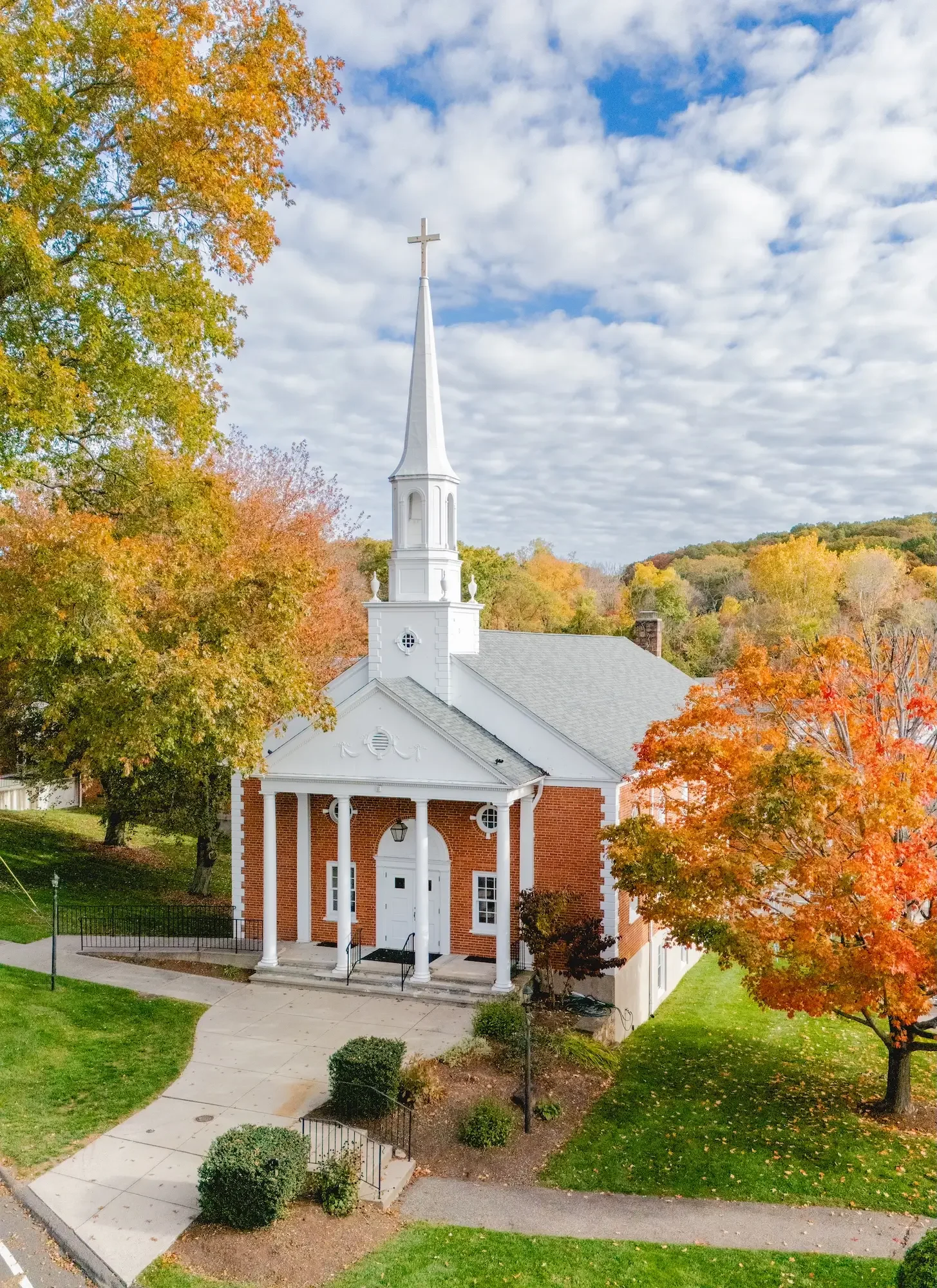 A white church with a steeple set among fall foliage with orange, yellow, and green trees and a partly cloudy sky.