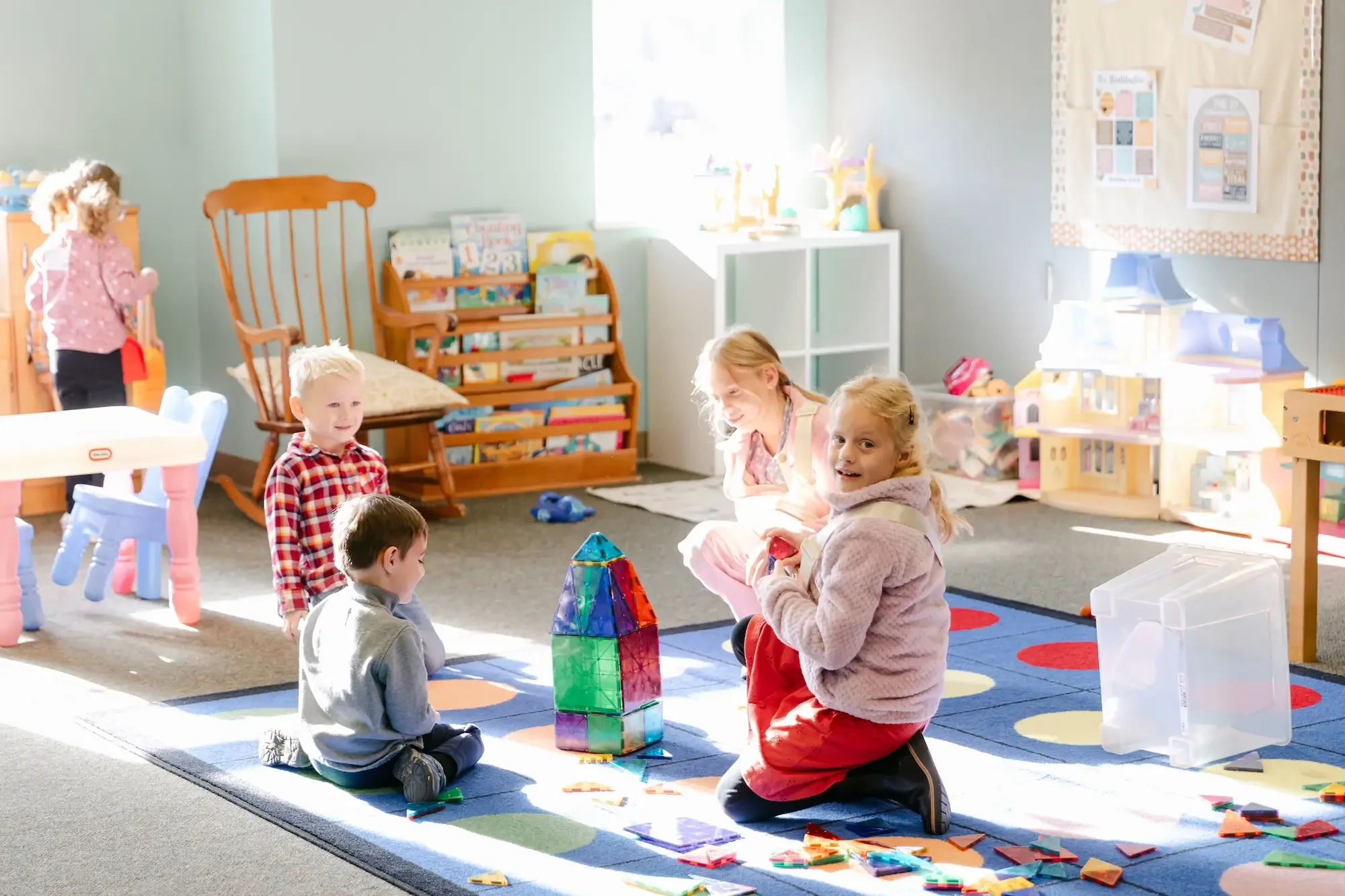Children playing with colorful construction toys and building a tower in a brightly lit classroom, surrounded by educational materials and furniture.