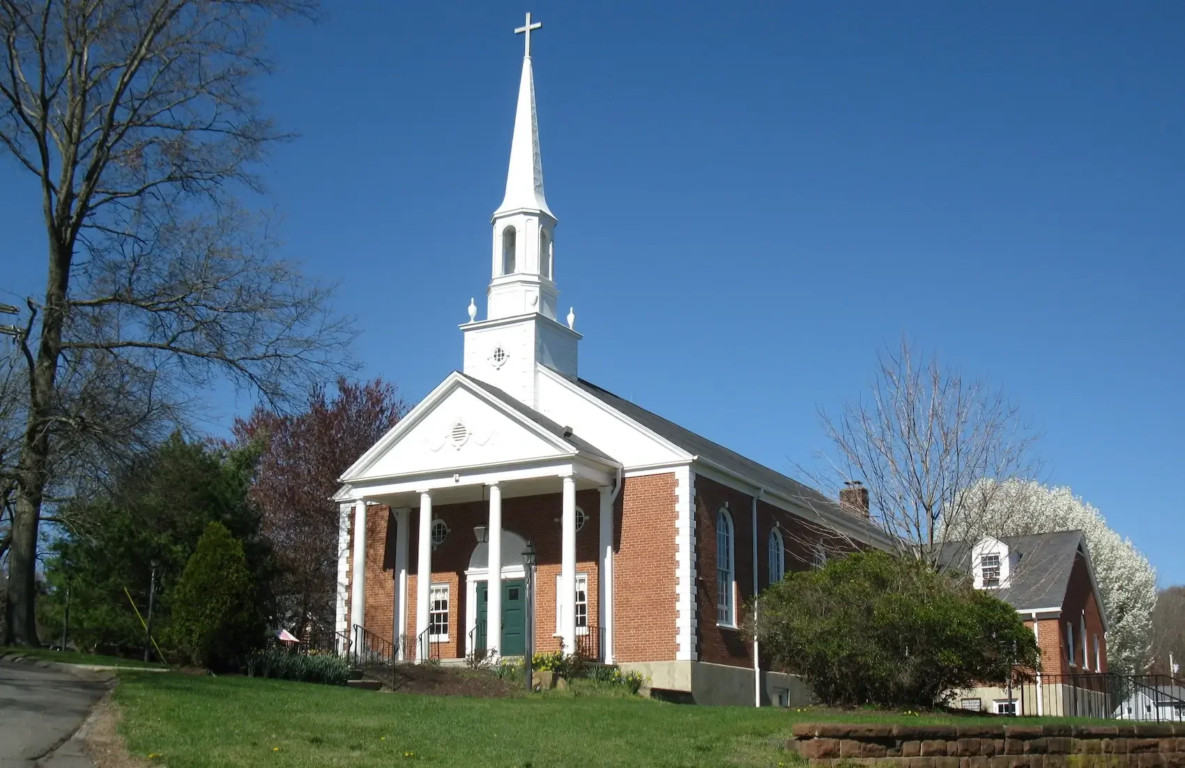 A white church with a tall steeple and brick walls, surrounded by trees and a grassy lawn, under a clear blue sky.