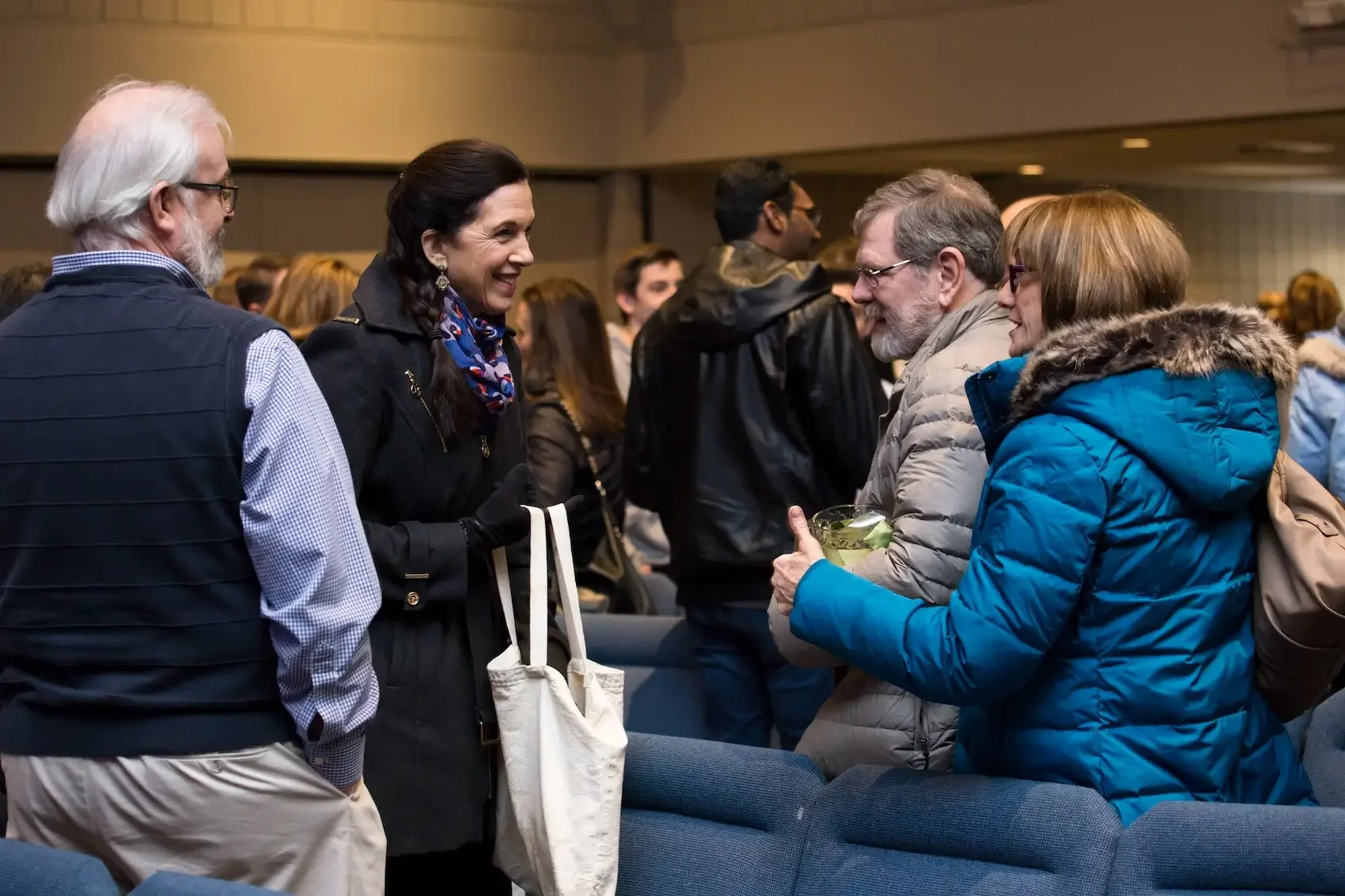 People engaging in conversation at an indoor event, with some holding drinks and wearing winter clothing.