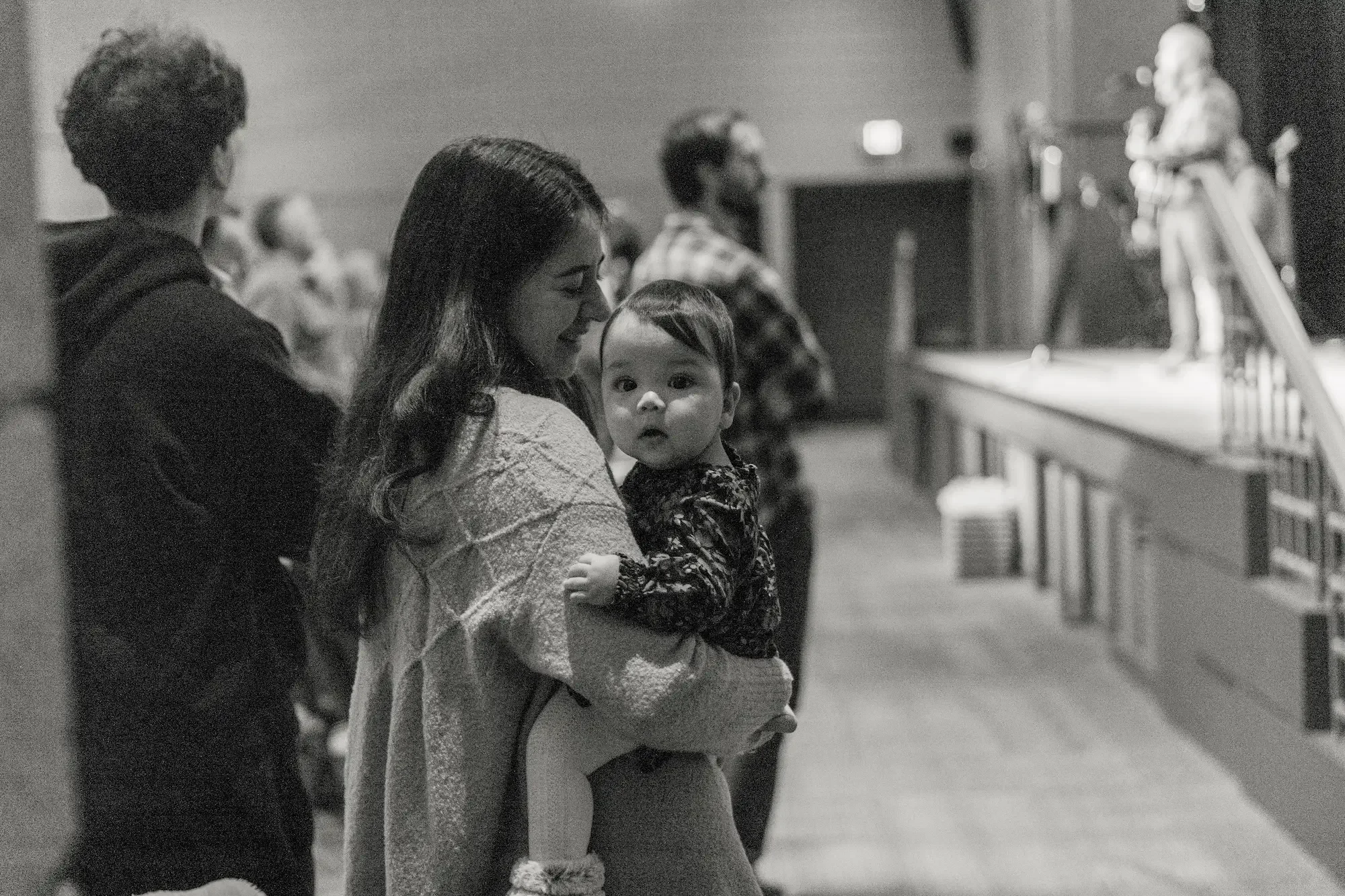 A woman holding a young child looking at the camera in a line of people at an indoor event, with a speaker on stage in the background.